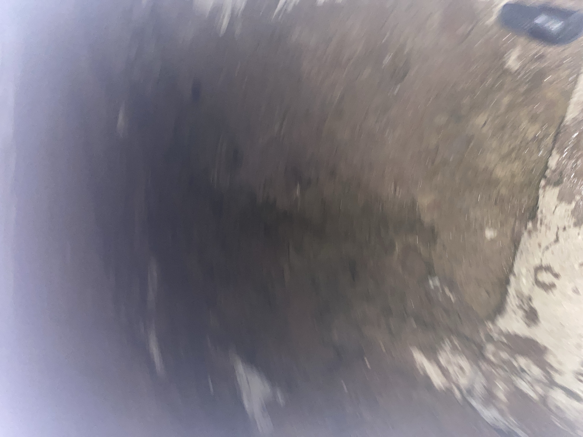 The image depicts a blurry, low-angle view of what appears to be a street or outdoor area in Jauro Yinu, Nigeria. The ground is wet and reflective, suggesting recent rain. There are patches of lighter-colored material, possibly debris or pavement, interspersed with darker, wet areas. In the lower right corner, a dark object, possibly a shoe or a small bag, is visible. The lighting is dim, and the overall impression is of a somewhat gritty, urban environment. The blurriness of the image may be due to motion or low light conditions.