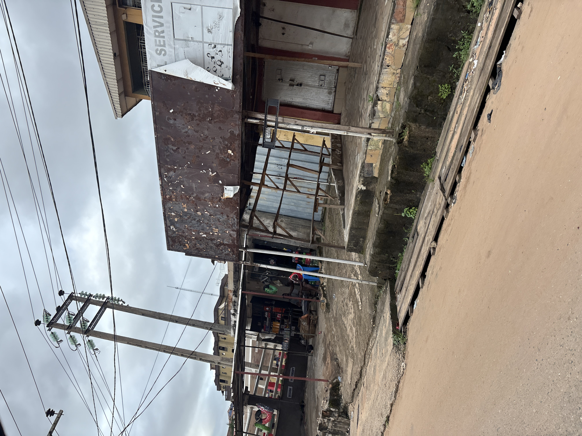 This image depicts a street scene in Nkpor, Nigeria, under an overcast sky. The foreground is dominated by a reddish-brown, dusty road. To the left, a concrete sidewalk with a slight elevation runs parallel to the road, showing signs of wear and tear, with some vegetation growing on its edge. A wooden plank bridge, constructed over a ditch, spans across the sidewalk and road.
In the midground, a series of stalls and shops are visible. One stall, covered by a corrugated metal roof supported by metal poles, displays various goods, including bottles and containers on shelves. Two men are seated in front of this stall; one is seated on a blue plastic chair, and the other is on a low stool. The stall appears to be a small shop selling household items or perhaps auto parts. Adjacent to this is a more substantial building with a weathered, rusty metal sign above it, which has the partially visible word "SERVICE" on it. This building has several entrances with closed doors.
In the background, there are more buildings and a prominent electrical pole with multiple wires attached to insulators. The overall impression is of a busy, commercial area in a developing urban environment, with evidence of daily life and commerce.