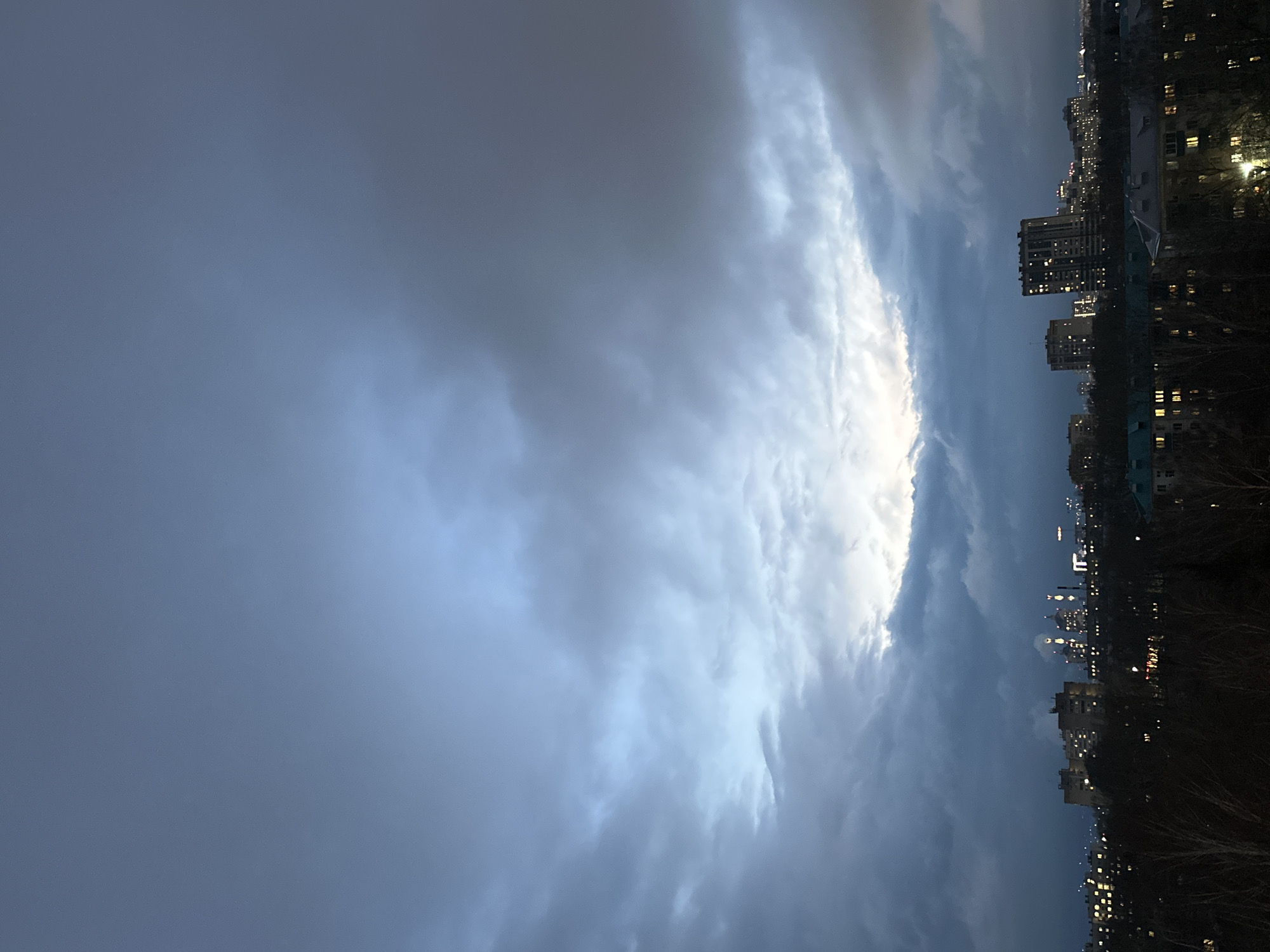 The image depicts a twilight cityscape in Kazan, Russia, under an overcast sky. Tall apartment buildings with illuminated windows line the right side of the frame, contrasting with the dark outlines of bare trees in the foreground. The sky is dominated by heavy, grey clouds, with a sliver of bright, white light visible between the cloud formations. The perspective suggests the photo was taken from a high vantage point.
