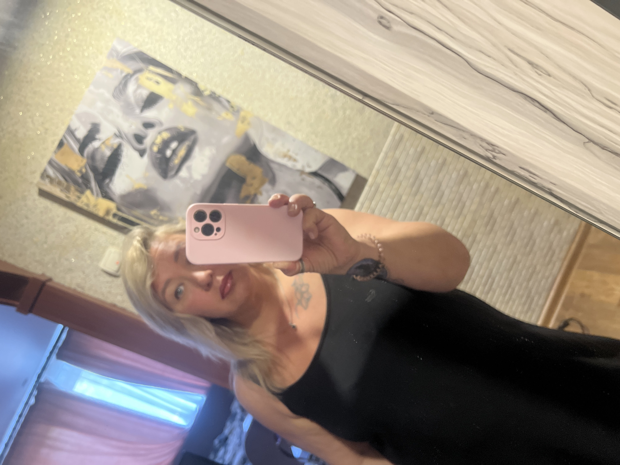 A fair-skinned woman with blonde hair is taking a selfie in a mirror. She is wearing a black tank top and jewelry on her wrist and neck. The reflection shows a wall with a black and white abstract art piece. The environment includes a partially visible window with curtains. The location cannot be determined as Chaikovsky, Russia.