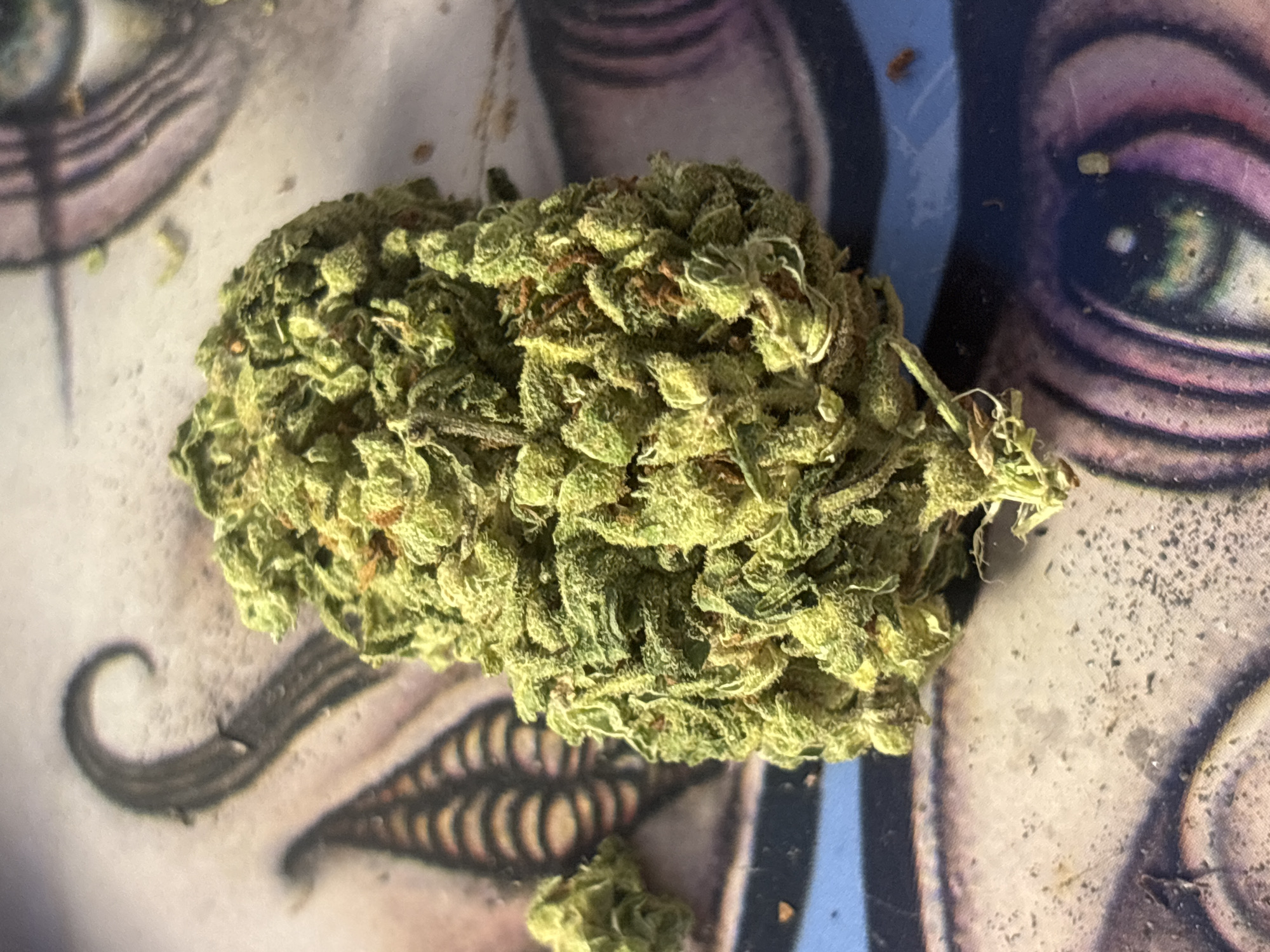 The image presents a close-up, top-down view of a large, dense cannabis bud positioned centrally on an illustrative surface. The bud is light to medium green, adorned with visible trichomes and interspersed with reddish-brown pistils, suggesting a mature and potent appearance. A smaller fragment of similar plant material is visible below the main bud.
The background surface features a multi-colored, detailed illustration, likely part of a rolling tray or mat. Prominent elements of this artwork include a stylized human eye with a green iris and black pupil on the right side of the frame, a curled, mustache-like design on the left, and a segmented, mouth-like pattern beneath the larger bud. The color palette of the illustration includes shades of blue, purple, black, and white.
No specific activity is taking place; the image is a still-life composition. The lighting appears artificial, illuminating the textures of the bud. Small particles and debris are visible on the surface surrounding the plant material. The time of day and weather are not discernible. While the location context is Buenos Aires, Argentina, the image itself does not contain any direct visual cues to confirm this. No text is visible within the image.
