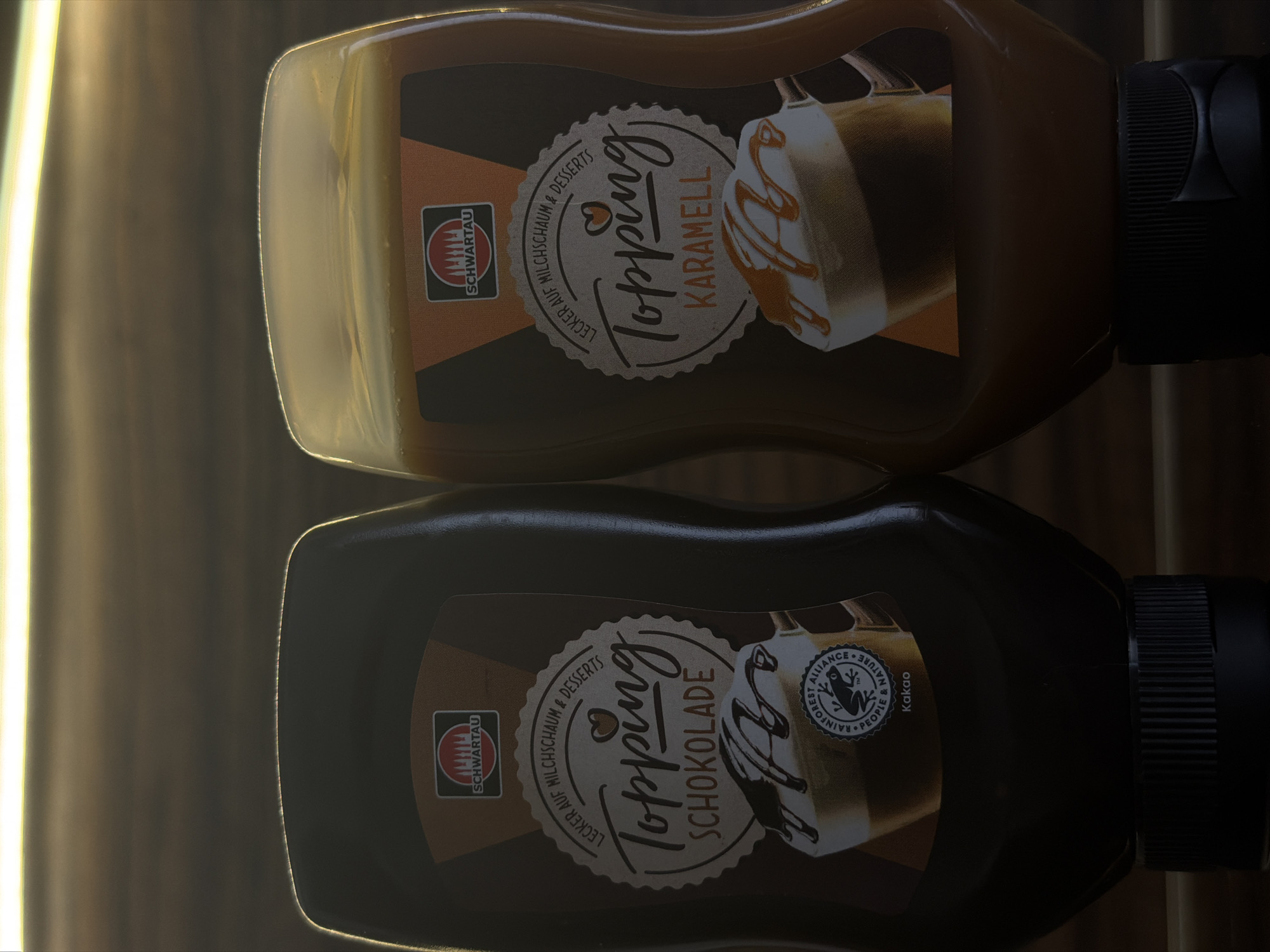 The image shows two bottles of Schwartau Topping syrups, one chocolate flavored and one caramel flavored. They are standing side-by-side on a dark, reflective surface, with a blurred background that suggests an indoor setting. The bottles are both clear plastic with labels. The chocolate topping bottle is on the left and the caramel topping bottle is on the right. The labels have a cream-colored circular design with text and images. The Schwartau logo, featuring red stylized trees within a red circle, is visible at the top of each label. The chocolate label says "Topping Schokolade" and depicts a cup of coffee with chocolate syrup. The caramel label says "Topping Karamell" and also shows a cup of coffee with caramel syrup. The image does not depict any people or actions. There is no information or visual evidence to suggest that this image is related to the city of Garmisch-Partenkirchen, Germany.