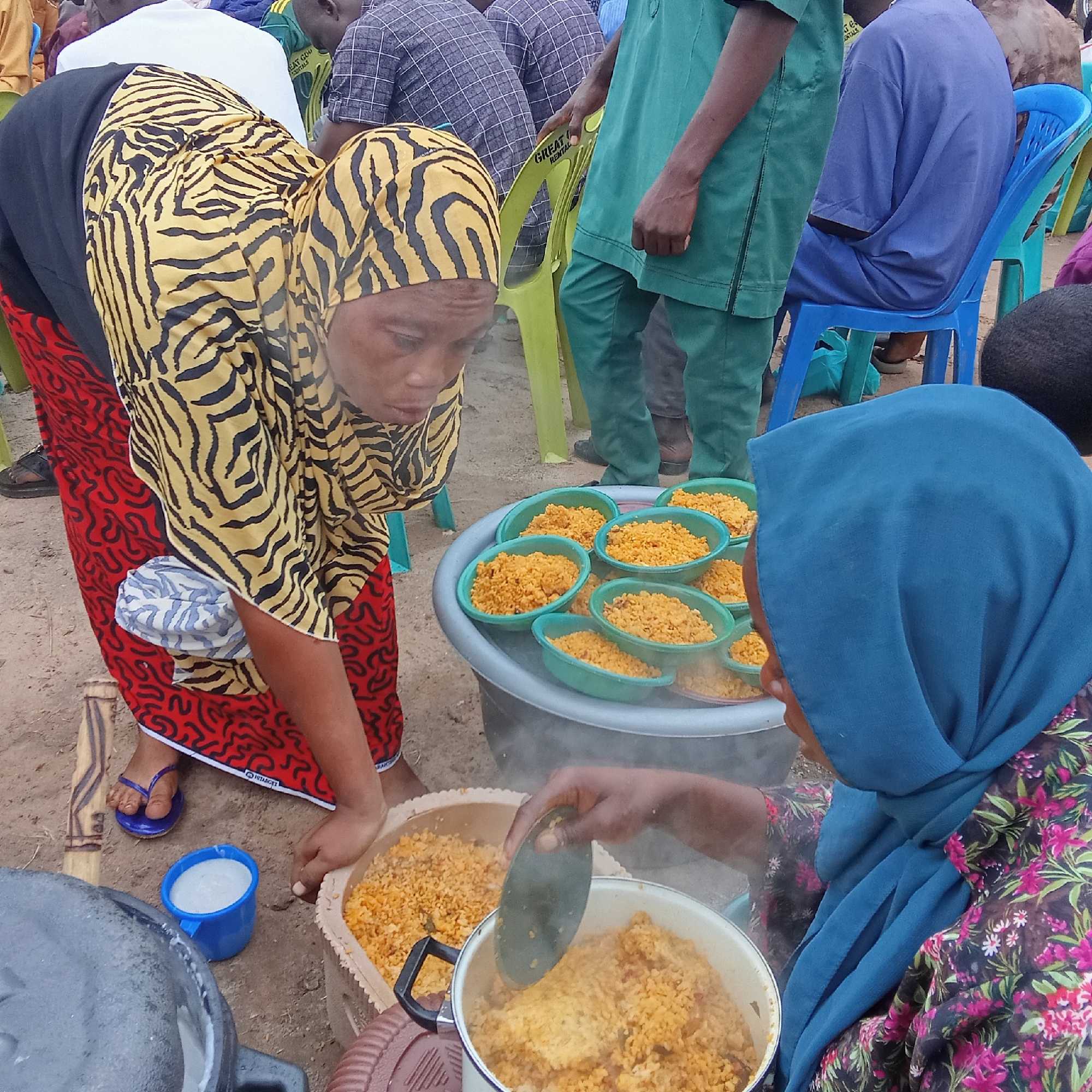 In Doma, Nigeria, two women are serving food. One woman, wearing a black and yellow zebra-striped headscarf, a black top, and a red patterned skirt, is reaching towards a container of cooked rice. Next to her is a blue plastic cup filled with a white liquid. The other woman, wearing a blue headscarf and a floral print top, is using a utensil to serve rice from a pot. Several green plastic bowls filled with rice are arranged on a large grey container. In the background, several people are sitting on green and blue plastic chairs. The ground appears to be sandy.