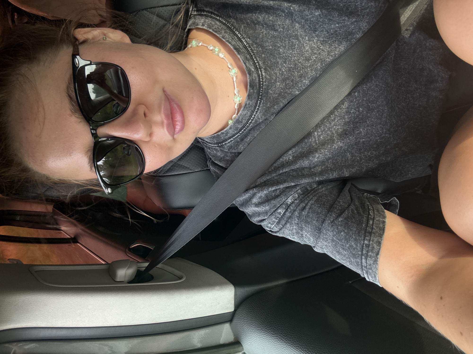 A medium shot captures a young woman with blonde hair pulled back, wearing black sunglasses, and a dark grey t-shirt with a light green flower-shaped beaded necklace. She is seated in a car, with a seatbelt visible across her chest. The interior of the car shows a gray seat and a white plastic trim piece. The window tint is a reddish-brown, and through it, greenery and trees are visible, suggesting a rural or suburban setting. The image does not contain any information about the city of Nylga, Russia.