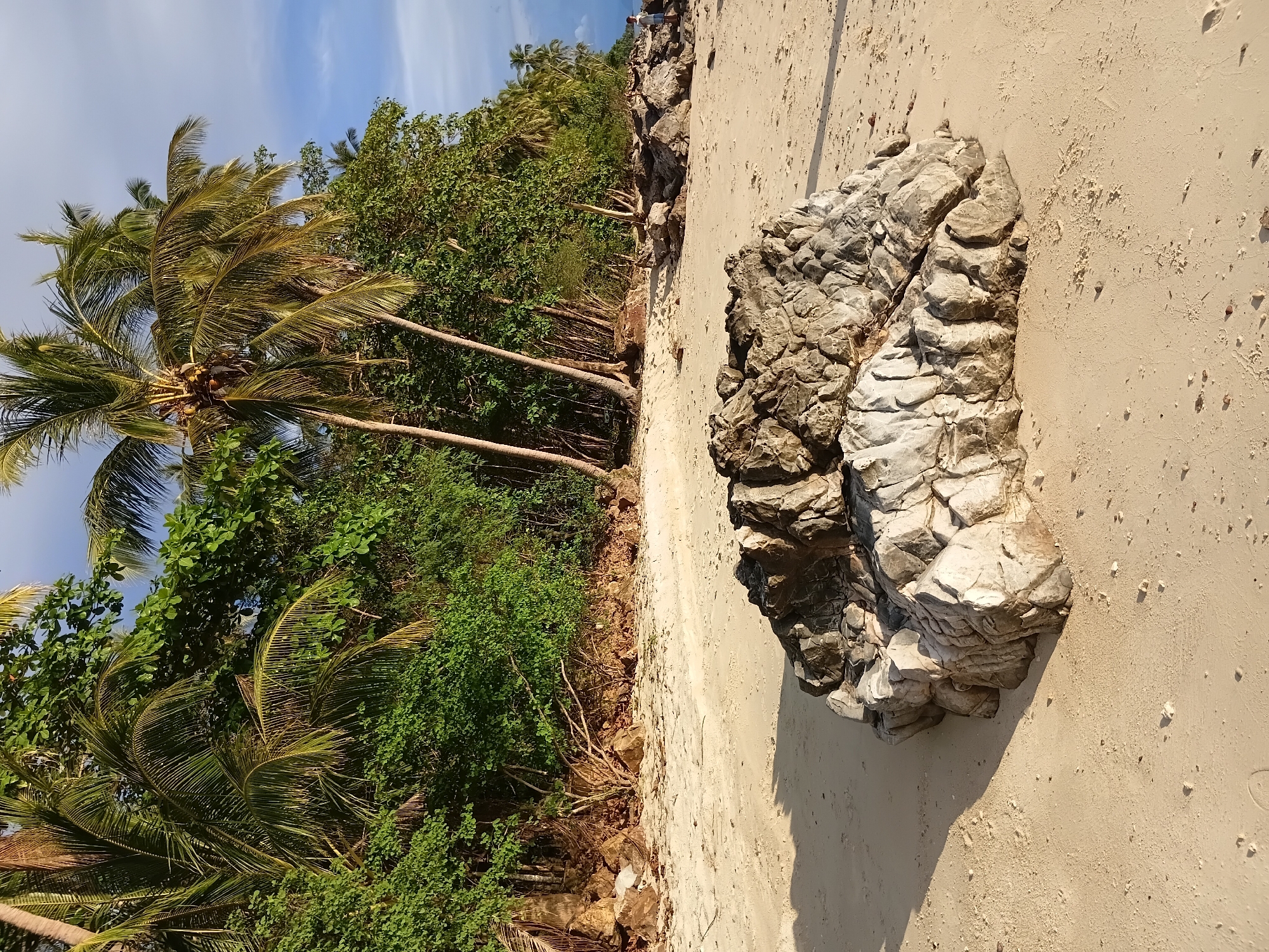 The image depicts a tranquil beach scene in Baan Pang Ka, Thailand. The foreground is dominated by a large, rugged rock formation situated on the sandy shore. Behind the rock, the sand stretches towards a dense line of lush green trees and palm trees, characteristic of a tropical environment. The sky above is a clear blue with a few wispy clouds, suggesting pleasant weather. The sunlight casts shadows across the sand, indicating it is likely late afternoon or early morning. No people are prominently featured in the immediate foreground, but a small figure is visible in the distance on the right side of the image, walking along the beach. The overall impression is one of a serene and natural coastal landscape.
