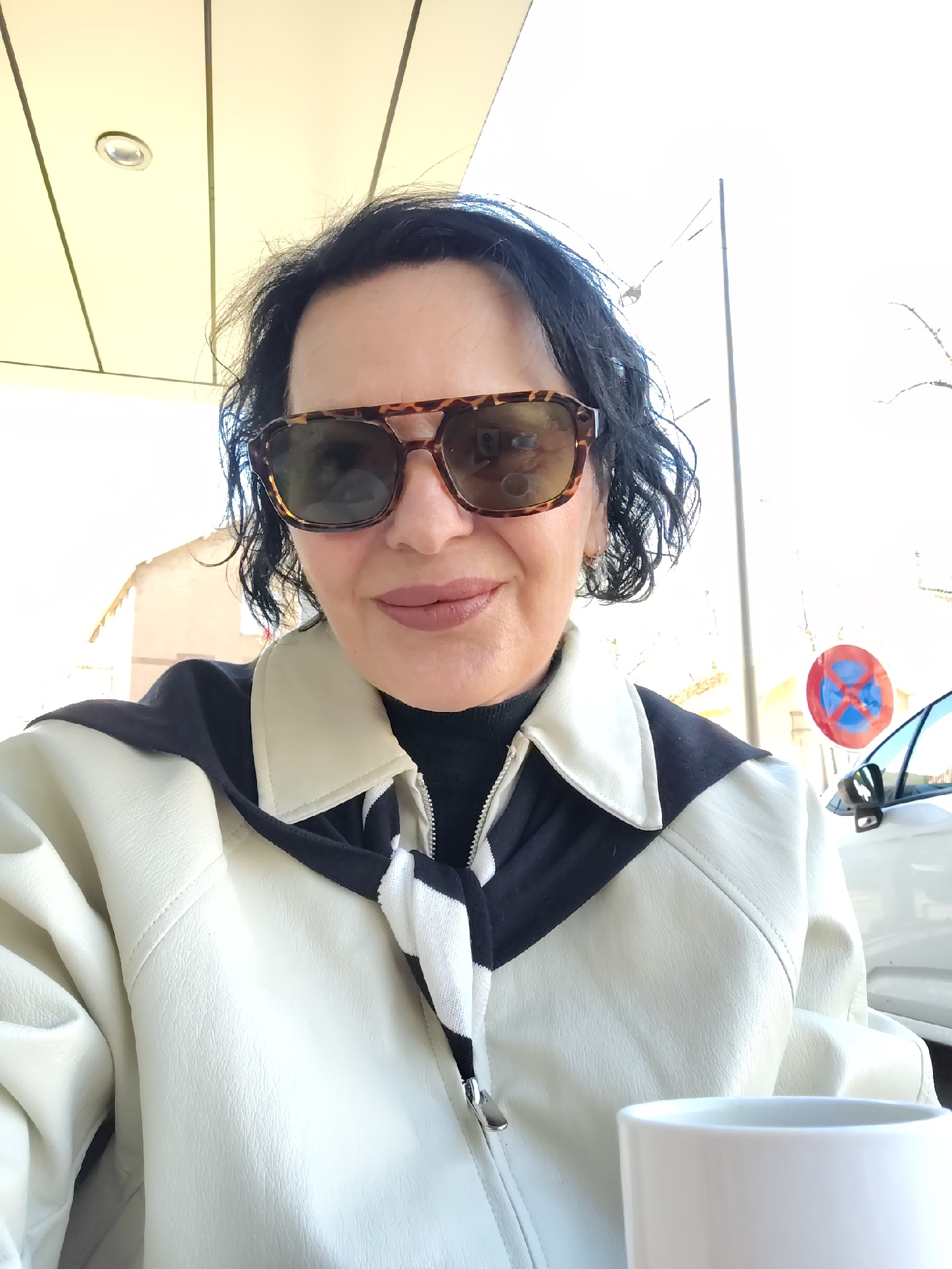 A woman is taking a selfie outdoors in Strasbourg, France. She is wearing sunglasses, a black turtleneck, and a light-colored jacket with black accents. She has short, dark hair and is smiling slightly. A white mug is visible in the lower right corner. A "no parking" sign is visible in the background, along with a white car. The location appears to be a cafe or outdoor seating area. The light suggests it is daytime.