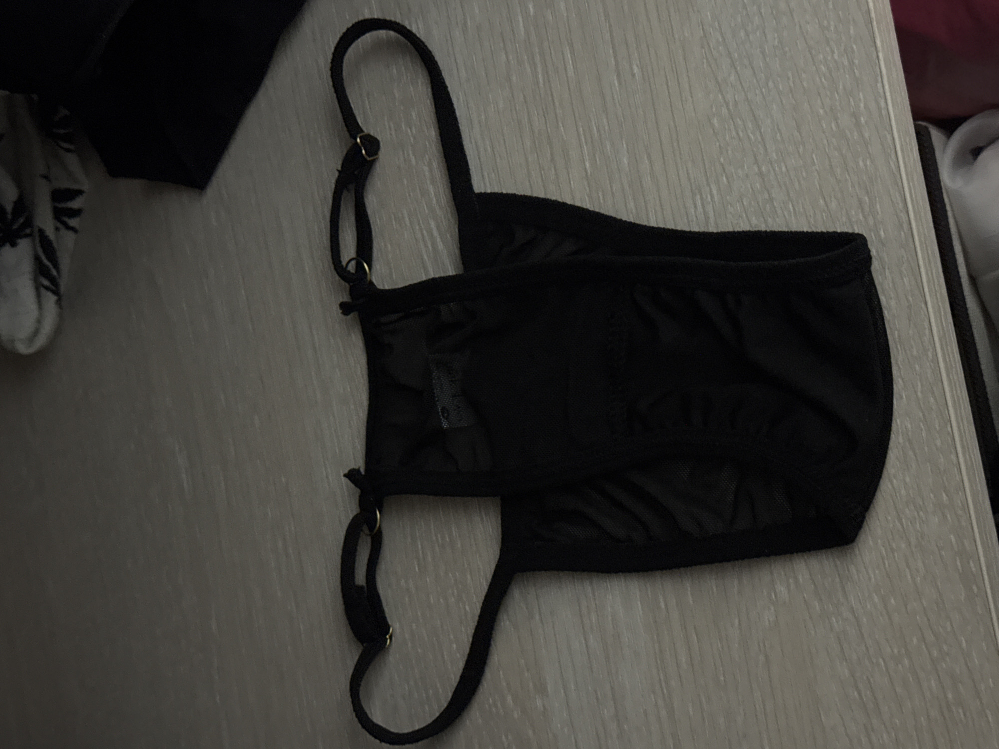 A black thong is laid flat on a light-colored, textured surface. The thong has thin straps with gold-colored adjusters. The fabric appears to be sheer in some areas. The background is out of focus and shows some other items of clothing. The image does not contain any information about the city of Saratov, Russia.