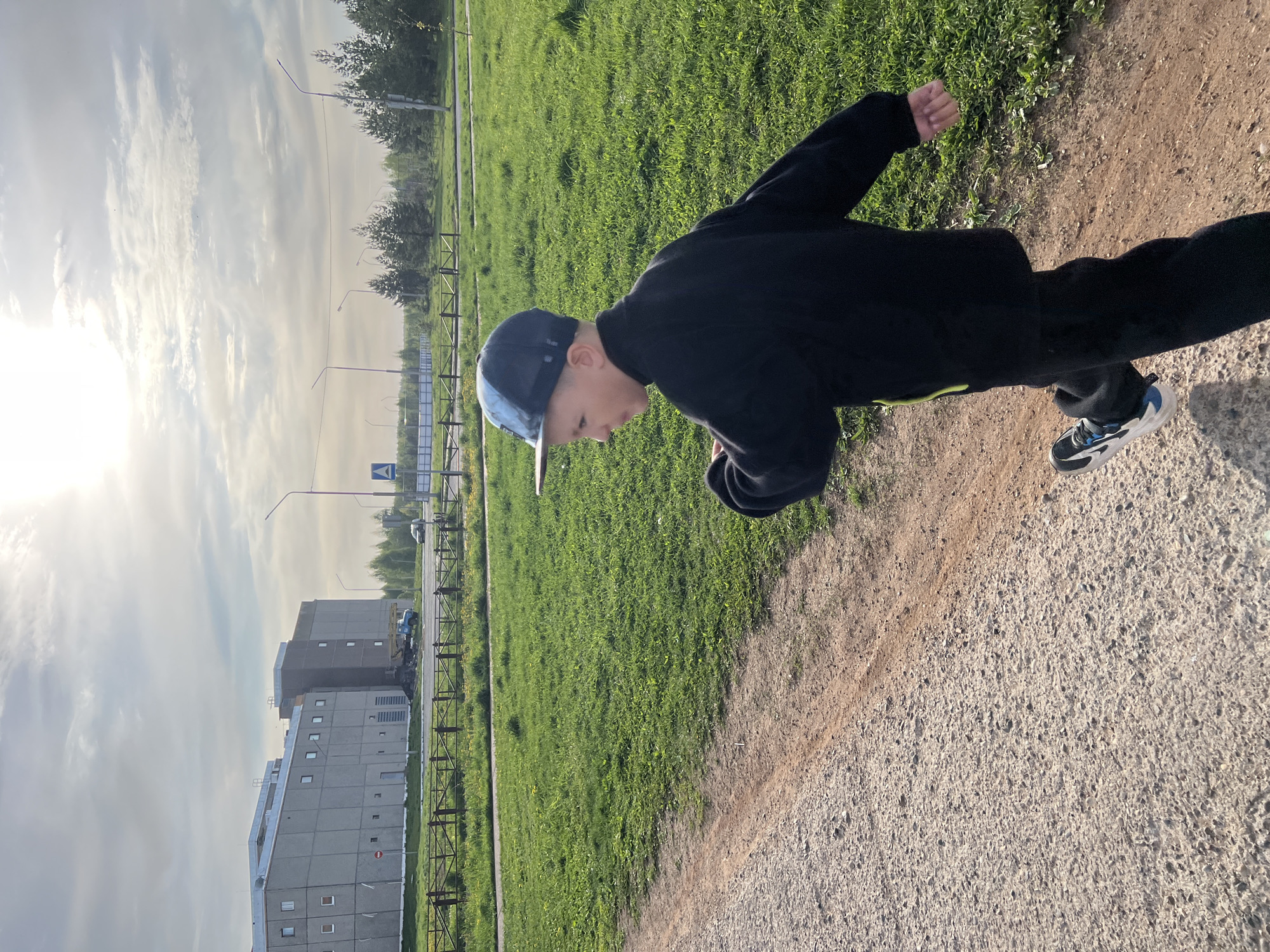 A young person wearing a black jacket and a baseball cap is walking on a dirt path. The path is adjacent to a grassy area. Behind the grassy area is a building and a street with a fence. The sky is cloudy. The location is Марковский, Russia.