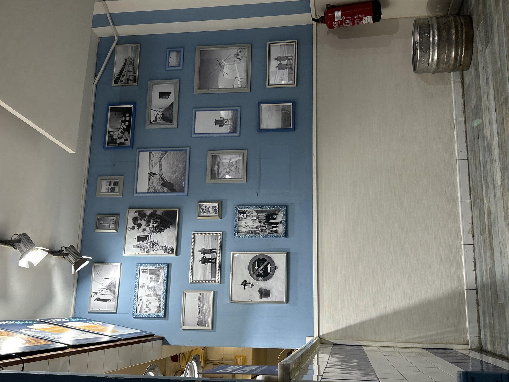 A vertically-oriented shot from within a building, possibly a restaurant or bar, displaying an eclectic collection of framed black-and-white pictures adorning a pale blue wall. The photos vary in size and style of frames, and in subject matter.
Above, a fire extinguisher is mounted on the wall, adjacent to a metal keg. Below, a row of adjustable spotlights illuminate a counter, over which food items are placed. The floor is tiled in a blue-and-white checkerboard pattern. A portion of a window can be seen in the bottom frame of the image.
Given the architectural style of the building, the wall decoration, and the tile patterns, a possible location could be a restaurant or bar in Barcelona, Spain. However, more details would be needed for a more precise pinpoint.