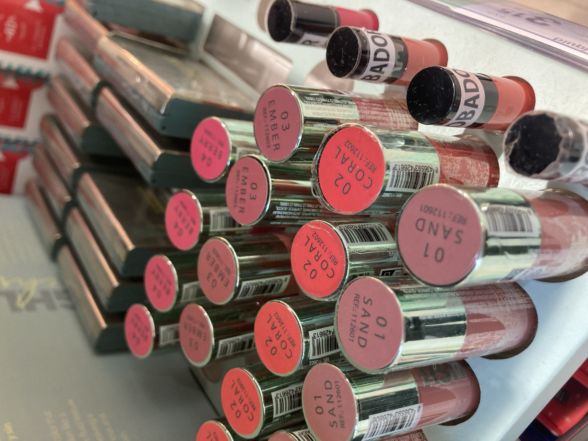 The image shows a collection of lipstick tubes, likely displayed for sale. The tubes are arranged in rows, with their colored tops facing outwards. The tops are labeled with numbers and names of shades, such as "04 BERRY," "03 EMBER," "02 CORAL," and "01 SAND." Some of the tubes have black caps with white text that appears to be a brand name, possibly "BADOH." The overall scene suggests a retail environment, perhaps a cosmetics store or a section of a larger store. The lighting is bright, indicating it might be daytime or under strong artificial lighting. There are no people visible in the image. The background is blurred but suggests more products or display elements. The setting is not definitively identifiable as Barcelona, Spain, but the provided context suggests this is the location. The overall mood is neutral, focusing on the product display.