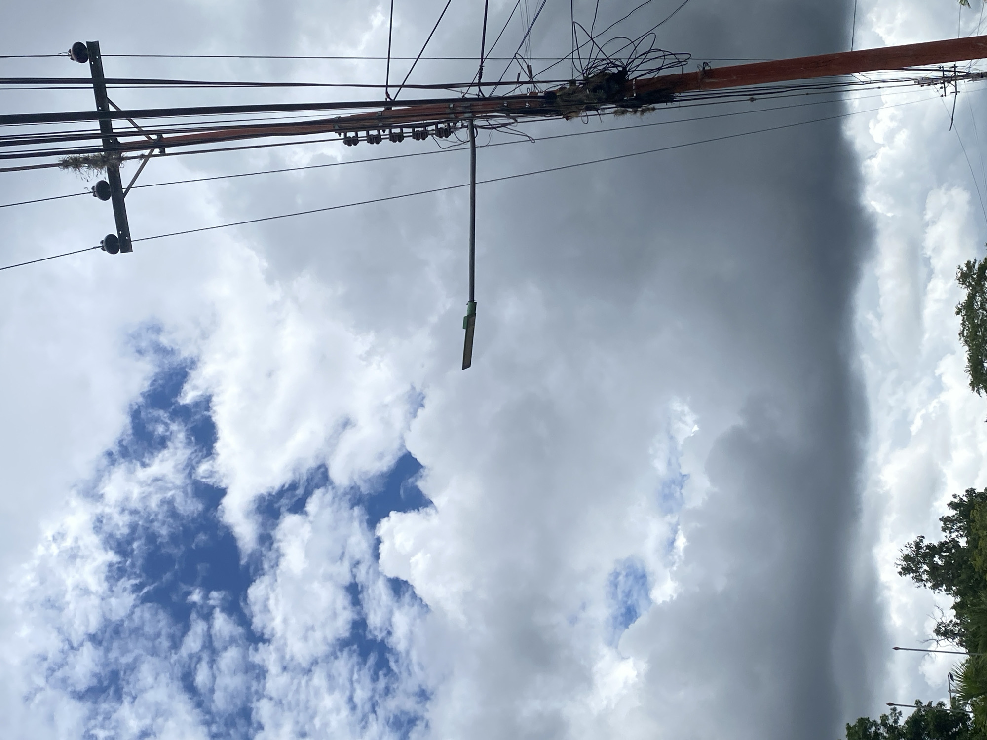 The image shows a view of the sky in Tinaquillo, Venezuela. The sky is filled with a mix of white, puffy cumulus clouds and darker, stormy-looking clouds. A blue sky is visible through some of the gaps in the clouds. Power lines and a utility pole run across the top of the frame. A street lamp is attached to the utility pole. Trees are visible in the bottom right corner of the image.