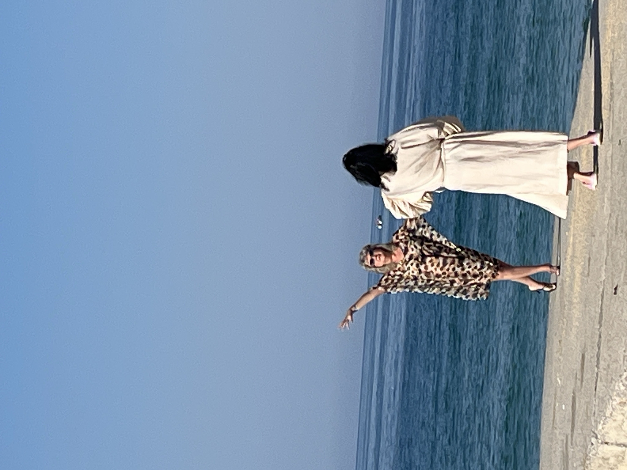 Two women are standing on a concrete pier or walkway next to the sea. The woman closer to the camera is wearing a patterned dress and sunglasses, with one arm outstretched. The other woman, further back, is wearing a long, light-colored robe or dress and pink sandals. The background shows a clear blue sky and the sea. The location is likely Alanya, Turkey.