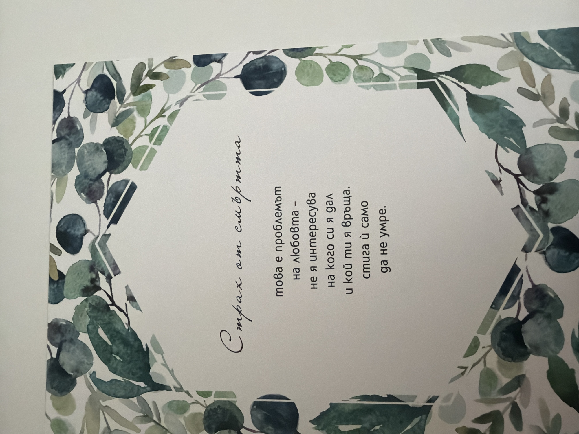 The image shows a close-up of a white card with a floral watercolor border in shades of green and blue. The text on the card is in Bulgarian and is written in a script font at the top and a standard font below. The top line of text appears to say "Страх от смъртта" (Fear of death). The lines below translate to: "This is a problem with love – / she is not interested / in who you are / and who you are around. / It is enough that you die." The image does not provide any information about a specific location in Pazardzhik, Bulgaria.
