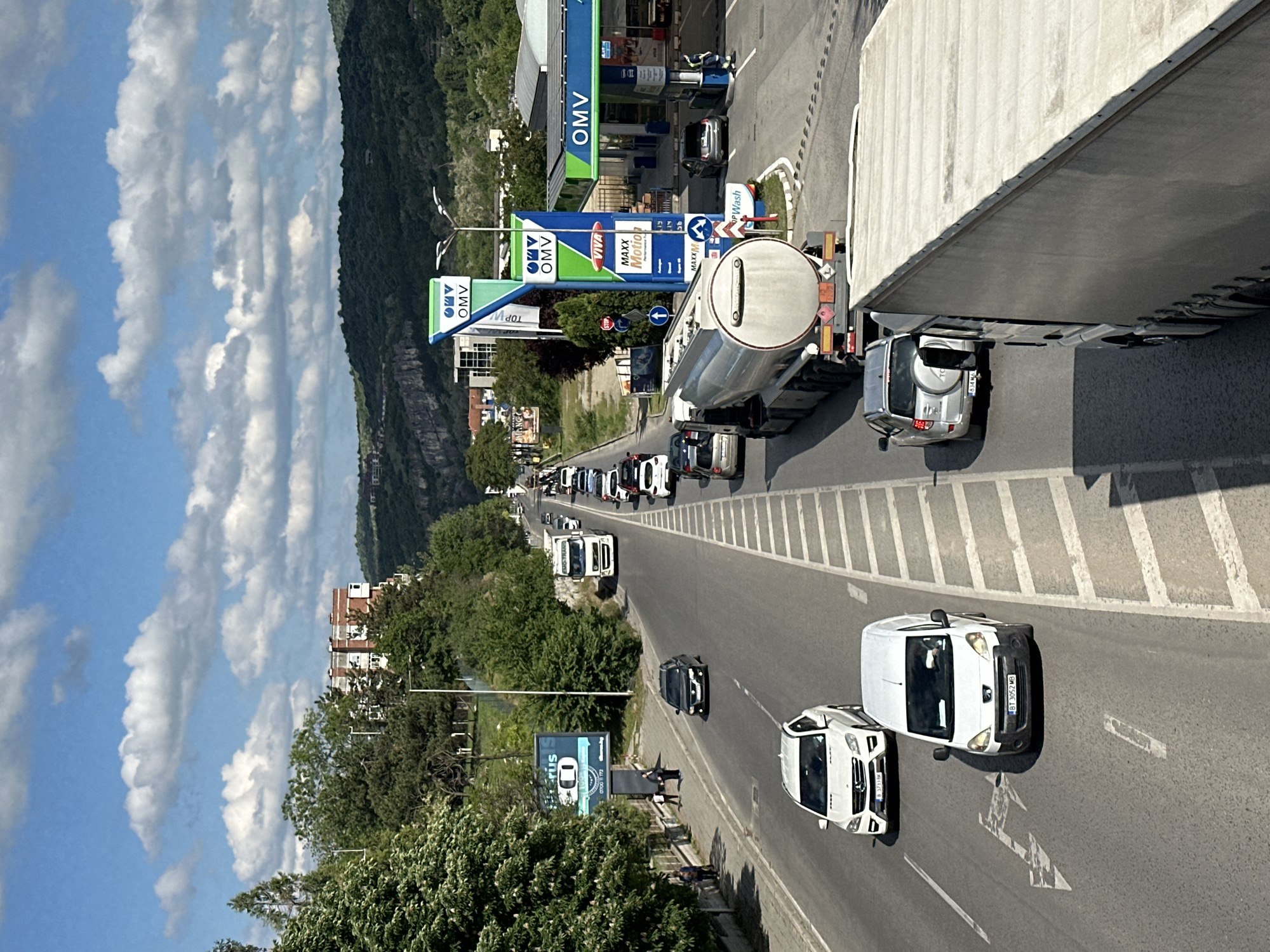 The image depicts a street scene in Veliko Tarnovo, Bulgaria. On the right side of the road, there is an OMV gas station. A tanker truck is delivering fuel to the station, causing a traffic backup. The sky is partly cloudy, and trees line the road. Several cars are visible, including a white van with the license plate "BT 3052 MB." A silver SUV partially obscured by a concrete structure has a license plate ending in "63446 O." The cars in the congestion have license plates "B3733BK" and "B3733KM". Pedestrians walk on the sidewalk next to a billboard advertising "Erus 0700 172." In the background, there is a mountain or hill.