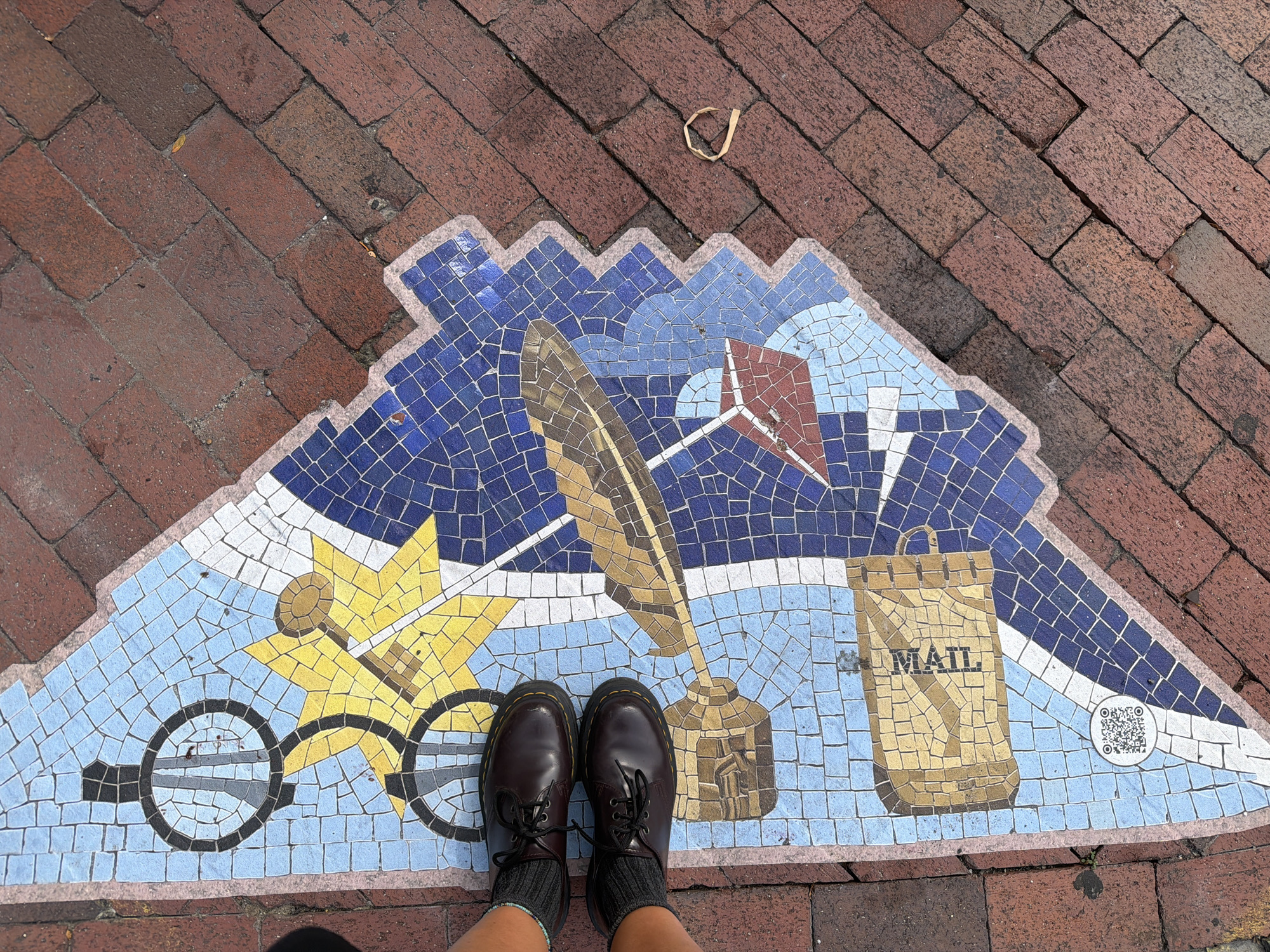 An overhead shot captures a section of a mosaic street art piece embedded in a brick-laid sidewalk in Philadelphia, Pennsylvania. The mosaic, in a fan shape, features various illustrated objects on a blue and white background. Prominently displayed are a yellow sunburst with a key, a quill pen, a mailbox with the word "MAIL" printed on it, and a QR code. Two pairs of feet wearing dark brown Dr. Martens shoes and grey socks are visible in the upper left corner, standing on the mosaic. The surrounding sidewalk is constructed of red bricks laid in a herringbone pattern. A small, dried leaf rests on the brickwork to the right of the mosaic. The lighting suggests it is daytime.