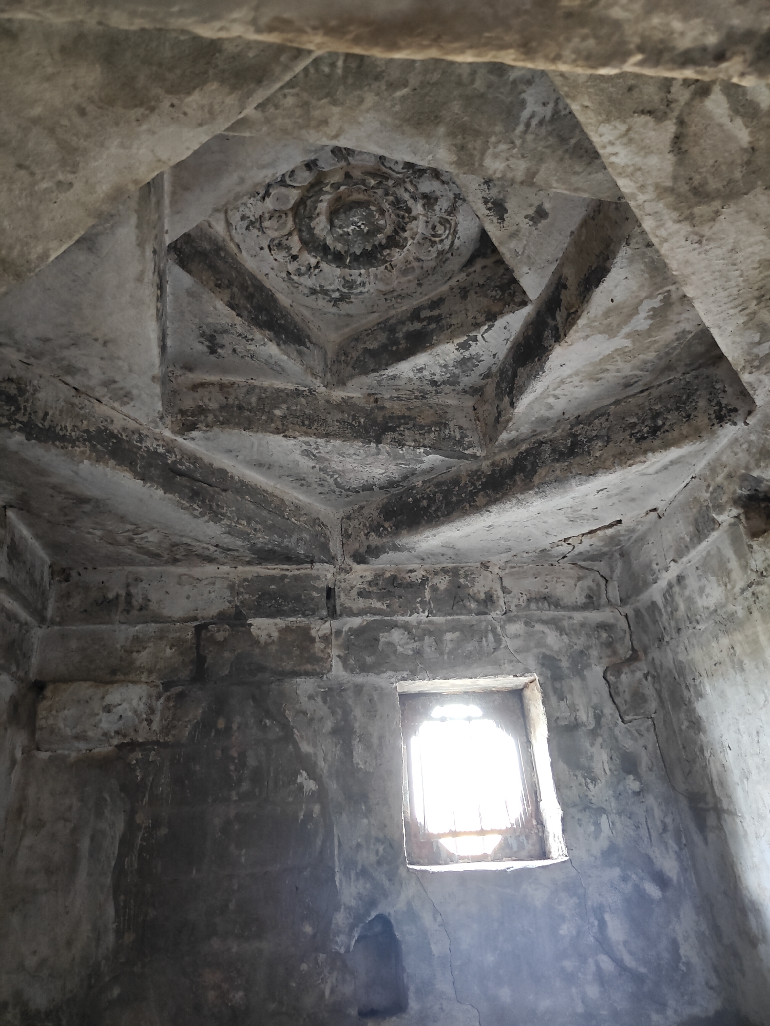 The photograph depicts the interior of a stone structure, likely a tomb or shrine, located in Kangra, India. The perspective is from the interior, looking upwards towards an elaborate, stepped ceiling. The ceiling is composed of stone slabs arranged in a geometric pattern, culminating in a central, circular, decorated rosette. The walls are constructed from roughly hewn stone blocks. A small, barred window is visible on one wall. The interior is dimly lit. The surfaces of the stone exhibit signs of age and weathering. There are no people visible in the frame. The general impression is one of age, decay, and architectural interest.