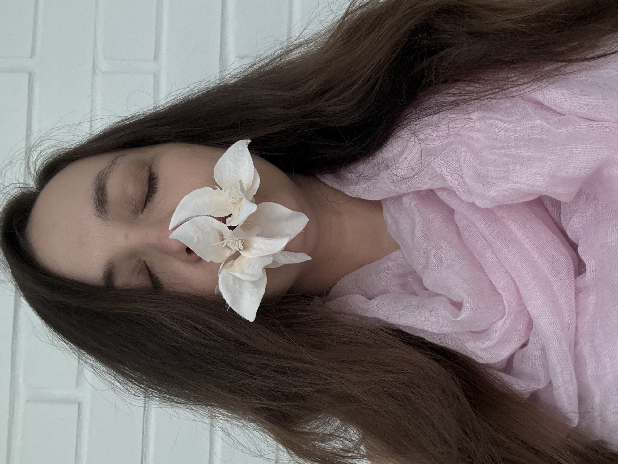 This vertical close-up photograph captures a serene moment featuring a person resting, possibly sleeping, with their eyes closed. The subject has long, flowing dark brown hair that spreads across the frame, framing their head and partially obscuring their left shoulder. A delicate white artificial flower, possibly an orchid or magnolia, with a slightly reddish-brown center, is gently placed over their nose and mouth, obscuring the lower half of their face.
The person's skin tone is fair, and their facial features, particularly the closed eyes and eyebrows, suggest a calm or peaceful state. Around their neck and upper chest on the right side of the image, a soft, light pink, sheer fabric, appearing to be a scarf or shawl, is draped in gentle folds, adding a touch of color and texture.
The background consists of a plain white, subtly textured surface, resembling painted brickwork or rectangular tiles, which provides a clean and minimalist setting for the portrait. The lighting appears soft and even, highlighting the textures of the hair, fabric, and flower. The overall mood conveyed is one of quietude, tranquility, and introspection. This scene is set in Chaikovsky, Russia. There is no visible text in the image.