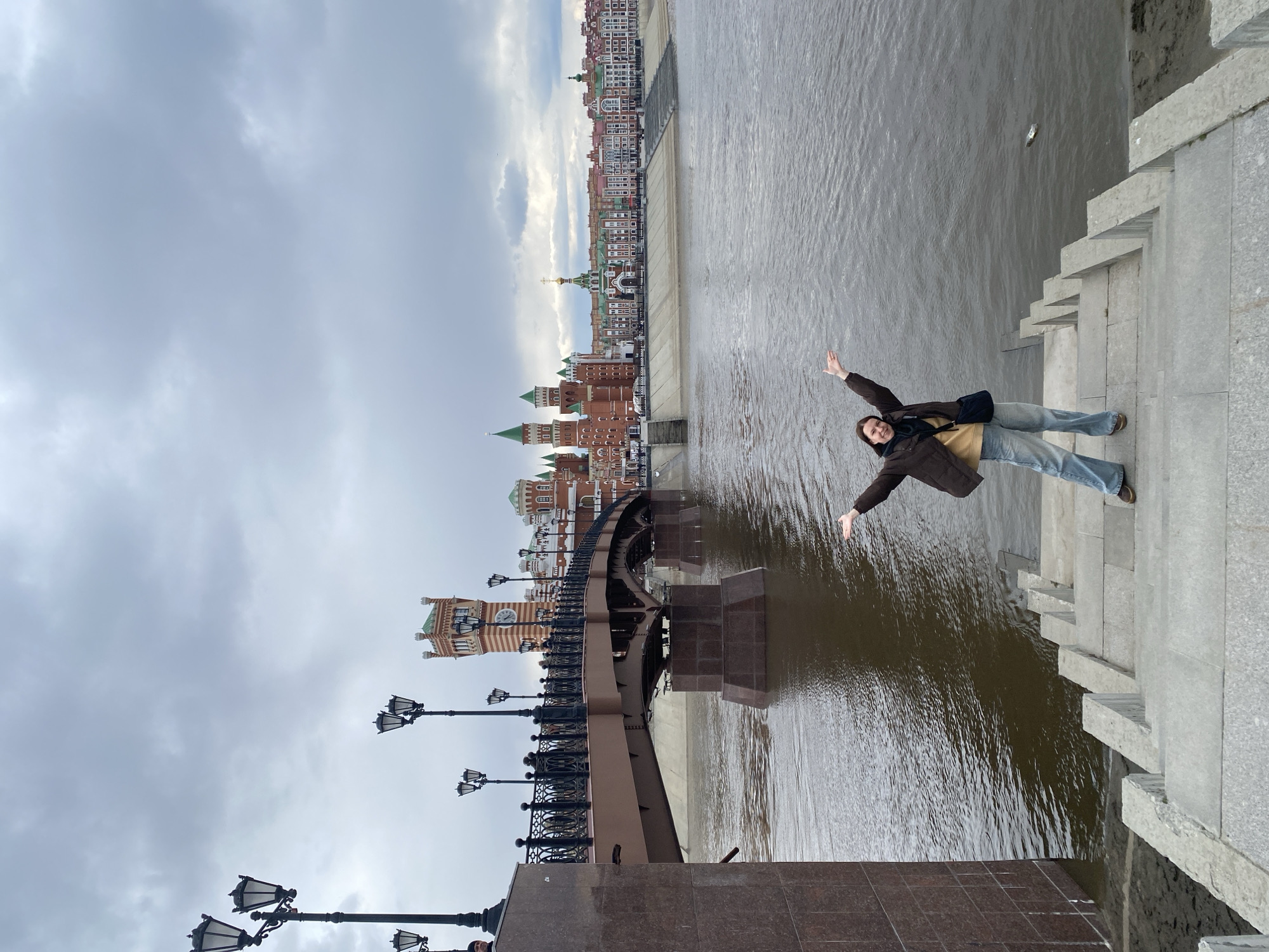 An outdoor, slightly rotated photograph captures a person striking a joyful pose on a concrete embankment overlooking a wide river in Yoshkar-Ola, Russia. The individual, an adult with short dark hair, is smiling broadly with both arms outstretched. They are dressed in a dark brown jacket, a light-colored top, and blue jeans.
To the person's left, a decorative metal bridge with ornate black lampposts stretches across the river. The bridge is supported by substantial granite piers. The water below is a wide, brownish-grey river with visible ripples.
The far bank of the river is lined with a distinctive cityscape. Numerous buildings feature red brick facades, green roofs, and various towers and spires, indicative of a European-inspired architectural style common in Yoshkar-Ola. A prominent building with a clock tower is visible near the bridge, and further down the bank, a structure resembling a church with a golden dome stands out. The sky above is overcast and grey with visible cloud formations. The overall impression is that of a cool, cloudy day. No text is visible in the image.