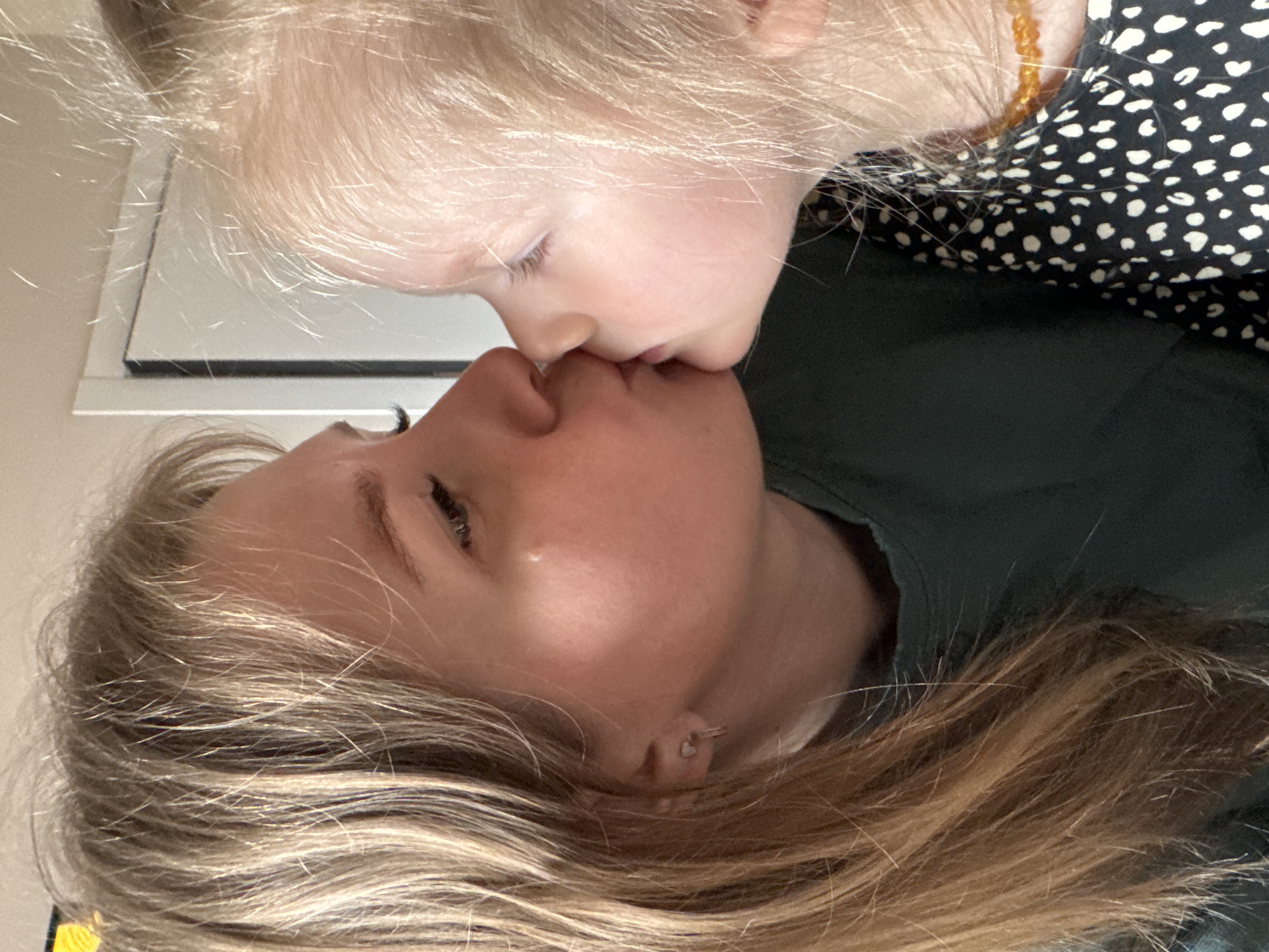 A fair-skinned woman and a fair-skinned toddler are positioned closely face to face, appearing to be kissing. The woman has blonde hair, visible eye makeup, and a small earring, and wears a dark green t-shirt. The toddler has light blonde hair, fair skin, and is wearing a black and white polka-dotted shirt and a beaded necklace. The background contains a light colored wall. The image gives no information about the city of Gornyi (Dvurechenskoe S/P), Russia.