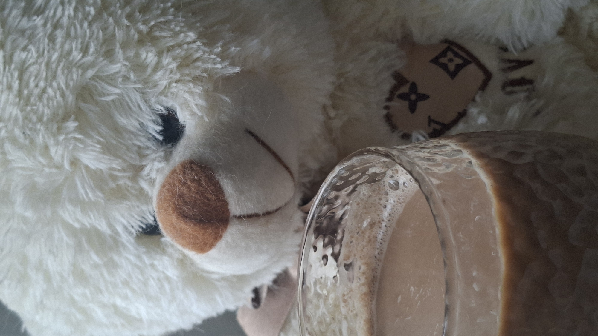 A close-up shot shows a white teddy bear with a brown nose and stitched smile next to a glass of foamy brown liquid, likely coffee or hot chocolate. The teddy bear has a heart-shaped patch on its chest with a repeating floral and diamond pattern and the letters "LV" visible within it. The background is soft and blurred, with the focus primarily on the bear and the drink. The scene appears to be indoors, possibly in a home setting, given the presence of a plush toy. There is no visible indication of the location being in Myadzyel, Belarus.