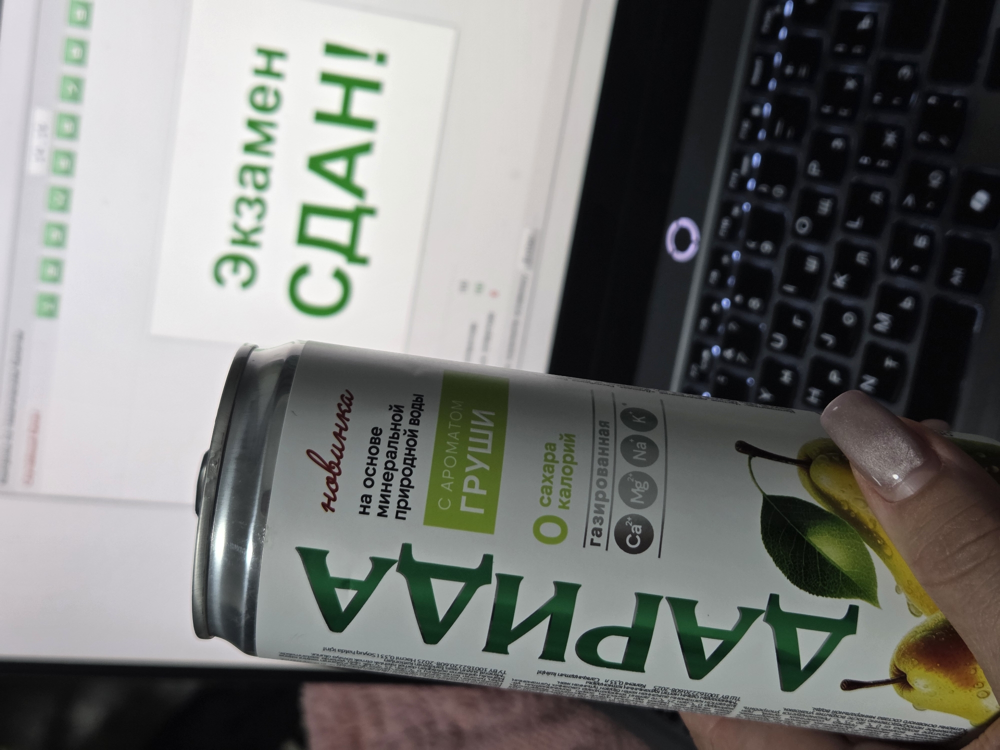 A person holds a can of "Darida" pear-flavored sparkling water in front of a laptop. The laptop screen displays the Russian text "Экзамен сдан!" which translates to "Exam passed!". The person's hand is visible, showing a polished fingernail. The laptop keyboard is partially visible. The background is blurry and out of focus. The location appears to be indoors.