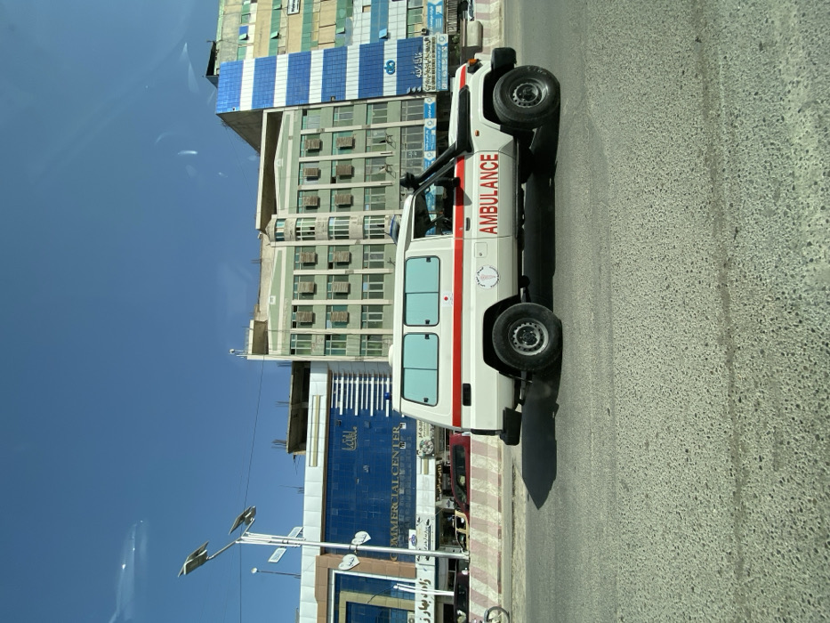 A white Toyota Land Cruiser J70 series ambulance, featuring a red horizontal stripe and "AMBULANCE" in red capital letters on its side, is positioned on an asphalt road. An emblem resembling the Afghan Red Crescent Society logo is visible on the vehicle's side, which also has light blue tinted windows and a snorkel on the passenger side. A blue emergency light is mounted on its roof. The ambulance casts a shadow to its left, indicating sunlight from the right. To the left of the ambulance, a concrete barrier with alternating red and white stripes separates the road from a sidewalk or parallel lane, where a partial bicycle wheel is visible.
In the background, under a clear blue sky, a series of multi-story buildings line the street in the city of Kandahar, Afghanistan. On the far left, a modern building with a blue reflective glass facade features signage including "COMMERCIAL CENTER" and text in Dari/Pashto script. A street lamp with two solar panels stands in front of this building. Adjacent to it, a light green/teal building with numerous windows and external air conditioning units rises several stories, with evidence of construction or renovation on its upper portion. Further to the right, another multi-story building displays a distinctive blue and white striped pattern on its facade.