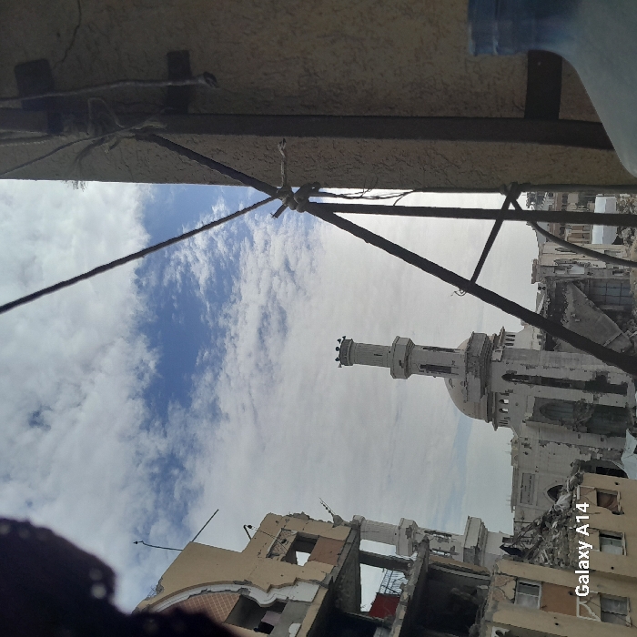 The image depicts a scene of extensive urban destruction in Khan Yunis, Palestinian Territory, under a partly cloudy sky. In the immediate foreground on the right, a textured beige wall or pillar is visible, to which several dark wires and cables are attached, some secured with white string. A portion of a large blue plastic bottle is present in the bottom right. On the left, a multi-story building with a light brown facade shows severe structural damage, with large sections of its walls collapsed, exposing interior rooms, rebar, and creating significant piles of rubble at its base. Centrally positioned in the midground is a mosque, characterized by a tall, white minaret with a crenellated top, on which several small dark objects (likely birds) are perched. A large white dome is partially visible behind the minaret. The mosque building itself exhibits considerable damage to its exterior walls, with areas of visible decay and accumulated debris around its foundation. Further in the background, additional structures appear to be heavily damaged or destroyed. The sky is predominantly light blue with scattered white cumulus clouds. No individuals are present in the visible scene. A "Galaxy A14" watermark is visible in the lower-left corner.