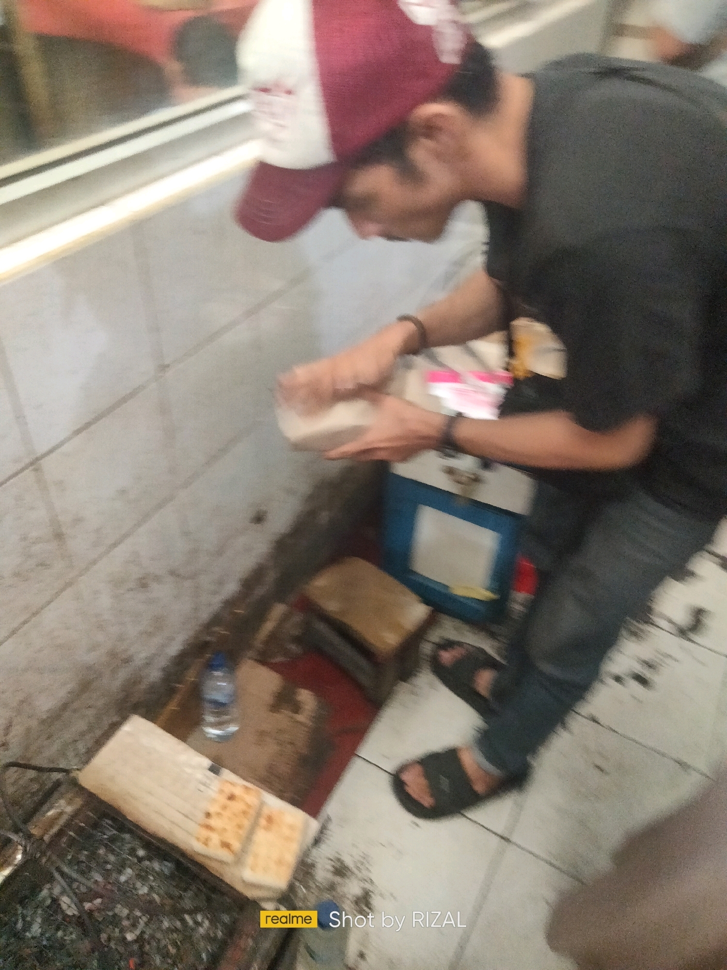 A person is seen wearing a black t-shirt, blue jeans, a maroon and white cap, and black sandals. They are holding a white paper container. The person is standing in front of a tiled wall and a light-colored floor. To the left, there's a grill with charcoal and what appears to be bread on top. A bottle of water is placed on a dark surface next to the grill. In the background, a blue cooler is visible. The scene is dimly lit and the image is somewhat blurry. The location is identified as Bojong Nangka, Indonesia.