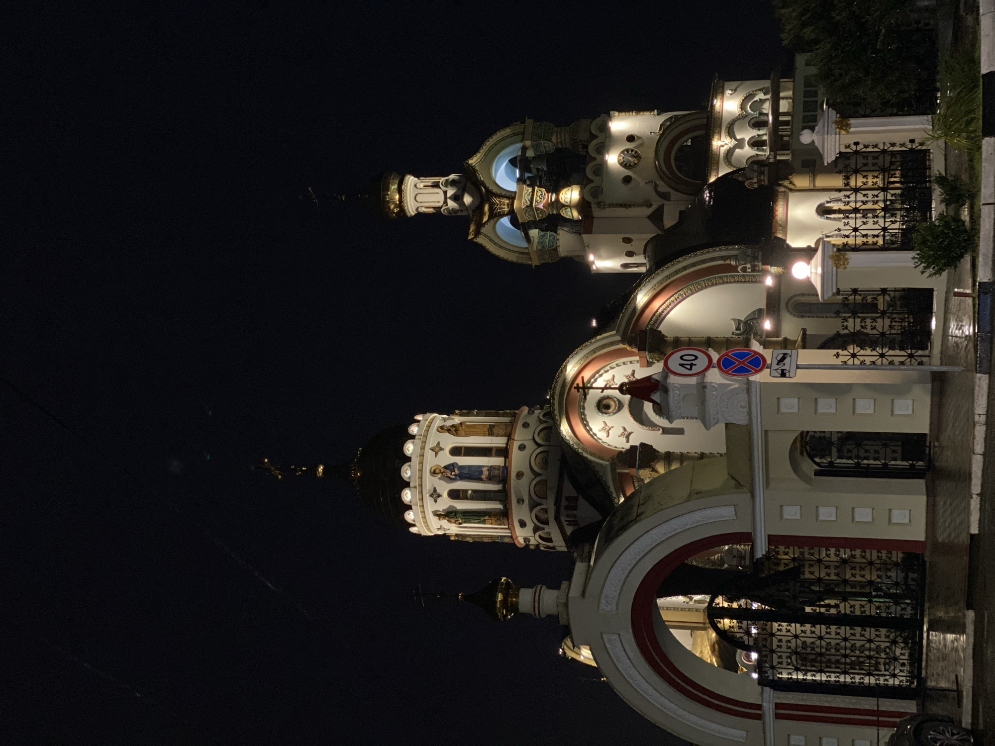 An eye-level outdoor shot presents a large Russian Orthodox church at night in Sochi, Russia. The primary focus is on the illuminated, ornate architecture, featuring domes of varying sizes and shapes, decorated with intricate patterns and religious iconography. The color palette is dominated by white and cream colors, highlighted by gold and red accents. A wrought iron fence runs along the base of the building, further emphasizing the architectural detail. Signage is visible near the building, with signs for traffic restrictions. The background is dark and mostly obscured, emphasizing the church as the central object. The street in front of the church is dimly lit, and a few details are visible of nearby vegetation and the street surface.