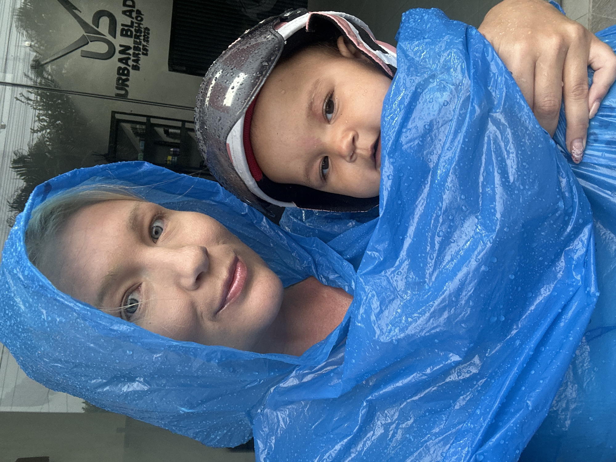 A woman and a child are huddled together under a blue plastic poncho. The woman is fair-skinned with blonde hair and blue eyes. The child is wearing a helmet. The background is blurred, but there appears to be a business storefront with the words "URBAN BLADE BARBERSHOP EST.2025" visible. The photograph appears to have been taken in North Kuta, Indonesia.
