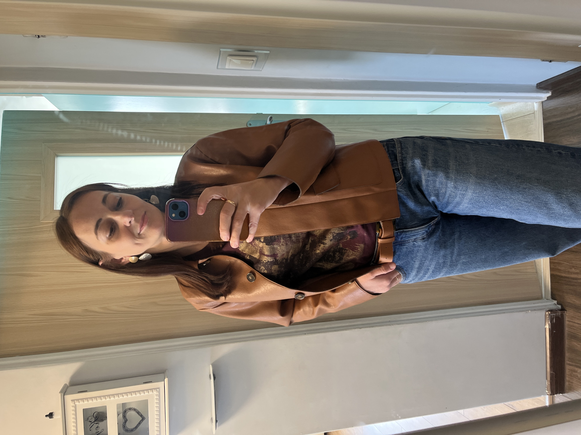 A woman with brown hair takes a mirror selfie in a room with wooden floors and light-colored walls. She is wearing a brown faux leather jacket over a patterned shirt and blue jeans. Her left hand is on her hip, and her right hand holds a phone. She is also wearing earrings and a ring on her right hand. A white cabinet with two framed pictures is visible on the wall to her left. The room appears to be indoors.