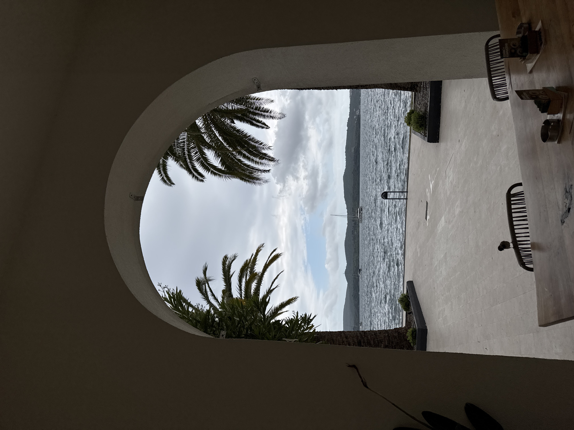 View through an arched opening showing a body of water, possibly the Bay of Kotor, in Tivat, Montenegro. The archway is part of a building, likely a restaurant or hotel, with palm trees partially obscuring the view. In the foreground, patio furniture suggests an outdoor dining area. The sky is overcast.