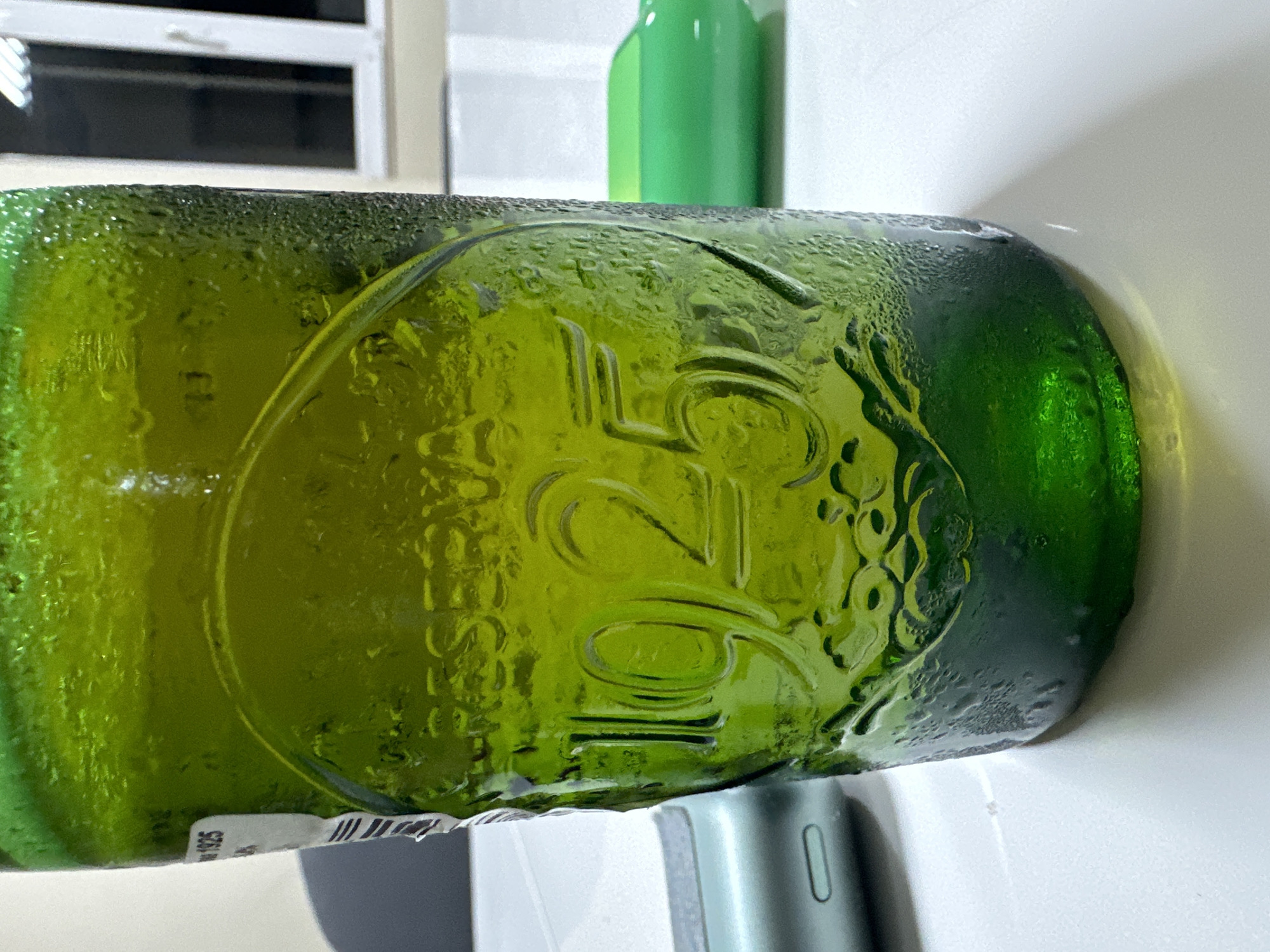 A close-up, horizontally oriented shot features a cold, green glass bottle covered in numerous condensation droplets. The bottle prominently displays a circular embossed design, which includes large numbers "125" or "12G" and a stylized letter "L" beneath the "12". To the right of these characters, further abstract patterns, possibly resembling grapes or a crest, are embossed into the glass. A small, partially visible white label at the bottom left of the bottle shows the number "1925".
The bottle rests on a white surface, with a dark grey and light blue/green electronic device, possibly an earbud case or vape, visible in the immediate foreground. In the background, another green bottle stands upright, and further back, a white window frame with dark interior panes is visible. The scene is set indoors, with the heavy condensation suggesting the main bottle is very cold, possibly recently removed from refrigeration. No specific activity is depicted. The location context provided for this image is Sofia, Bulgaria.