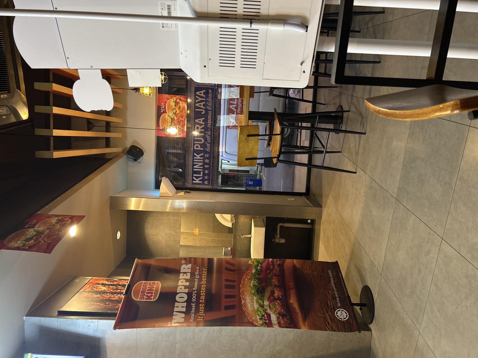 The image depicts the interior of a Burger King restaurant in what appears to be a densely populated urban area. Signage is visible outside, including what appears to be a medical clinic named "KLINIK PUTRA JAYA" with script in multiple languages including what appears to be Tamil or another Dravidian language. The restaurant's interior features modern design elements, including high-top seating with black frames. Burger King advertising materials, such as a "WHOPPER" banner stand are prominent. The restaurant is illuminated with recessed lighting and pendant fixtures. The location appears to be in the city of Subang Jaya City Council, Malaysia.