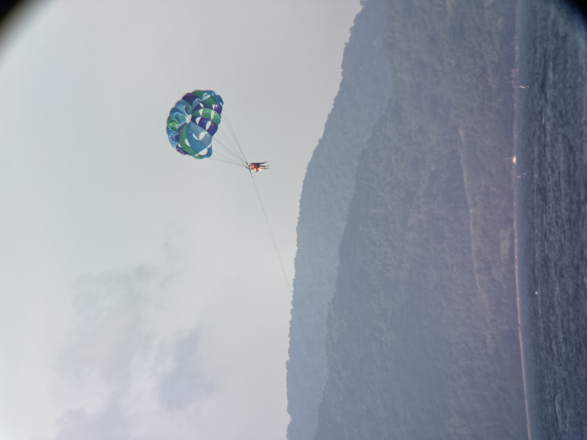 A parasail with two people is in the sky above the water in Da Nang, Vietnam. The parasail is colorful, with blue, green, and white sections. The two people are wearing life vests. Below them, a large, green, tree-covered mountain range stretches across the background. In the foreground, dark blue water is visible with some small white caps. The sky is overcast and a light gray.