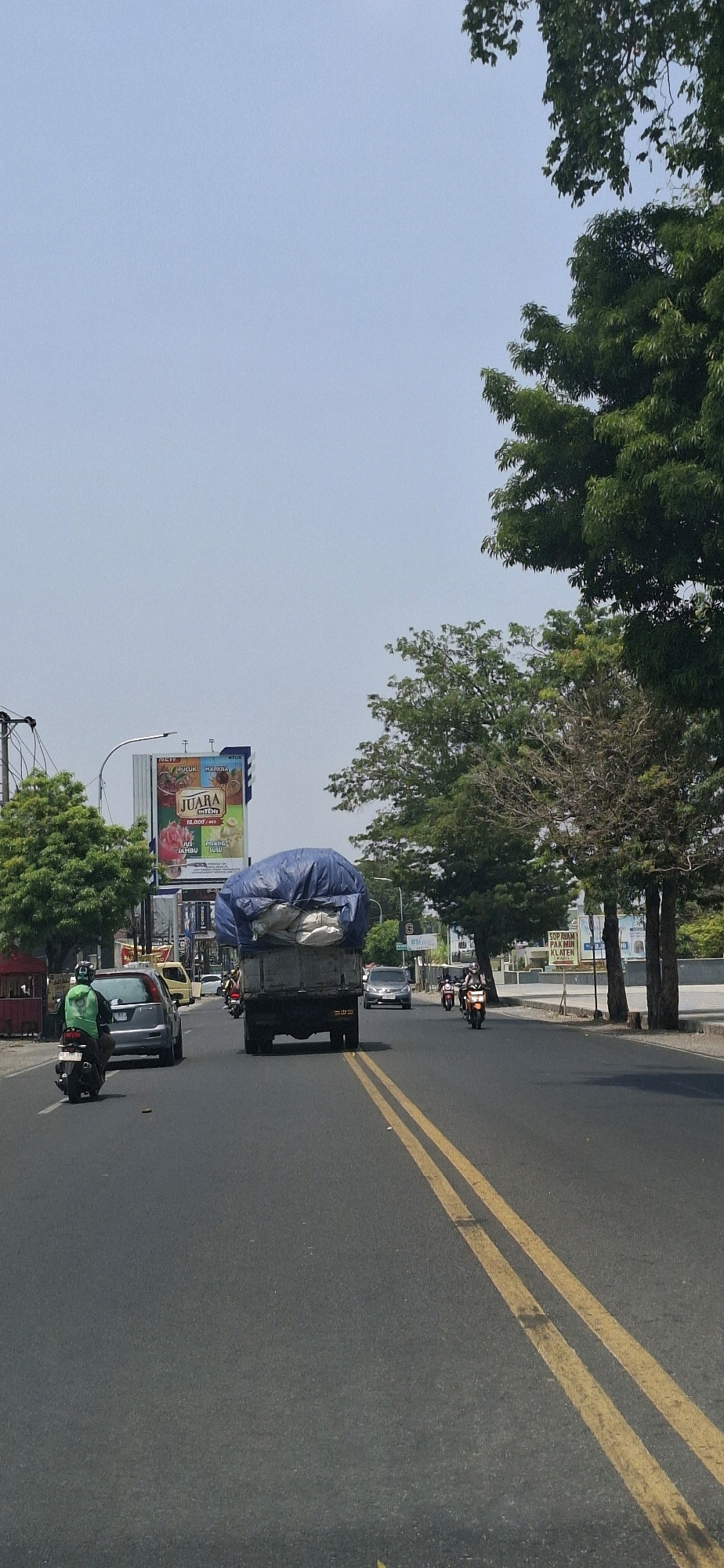 The image depicts a street scene in Colomadu, Indonesia, on a bright, clear day with a pale blue sky. The asphalt road features double yellow lines in the center, indicating a two-way traffic flow.
In the foreground, a person wearing a green jacket rides a motorcycle in the left lane, moving away from the viewer. To their right, a silver car is also in motion. Further ahead in the right lane is a large truck with its cargo covered by a blue tarp. The truck appears to be carrying a substantial load. Several other motorcycles are visible further down the road, also in motion.
The background reveals a cityscape with buildings and a large billboard displaying advertisements. Trees line the side of the road, some with full green foliage and others appearing more sparse. Streetlights and utility poles are also present. The overall impression is that of a busy urban environment.