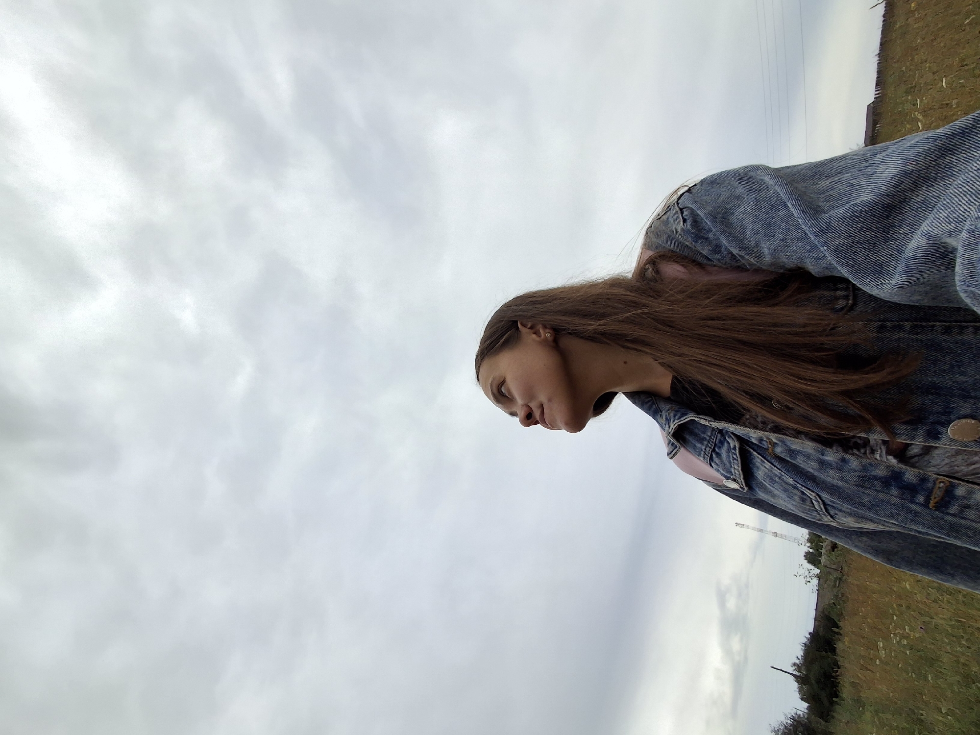A low-angle, eye-level medium shot captures a young Caucasian woman from the chest up, looking up at a cloudy sky. She has long, brown hair that falls past her shoulders. She is wearing a denim jacket over a black t-shirt. The jacket has pink accents on the shoulders. The background consists of a field of dry grass and some sparse trees on the left. In the distance, a cell tower is visible. The overall lighting is dim, suggesting an overcast day. The location is the city of Garevaya, Russia.
