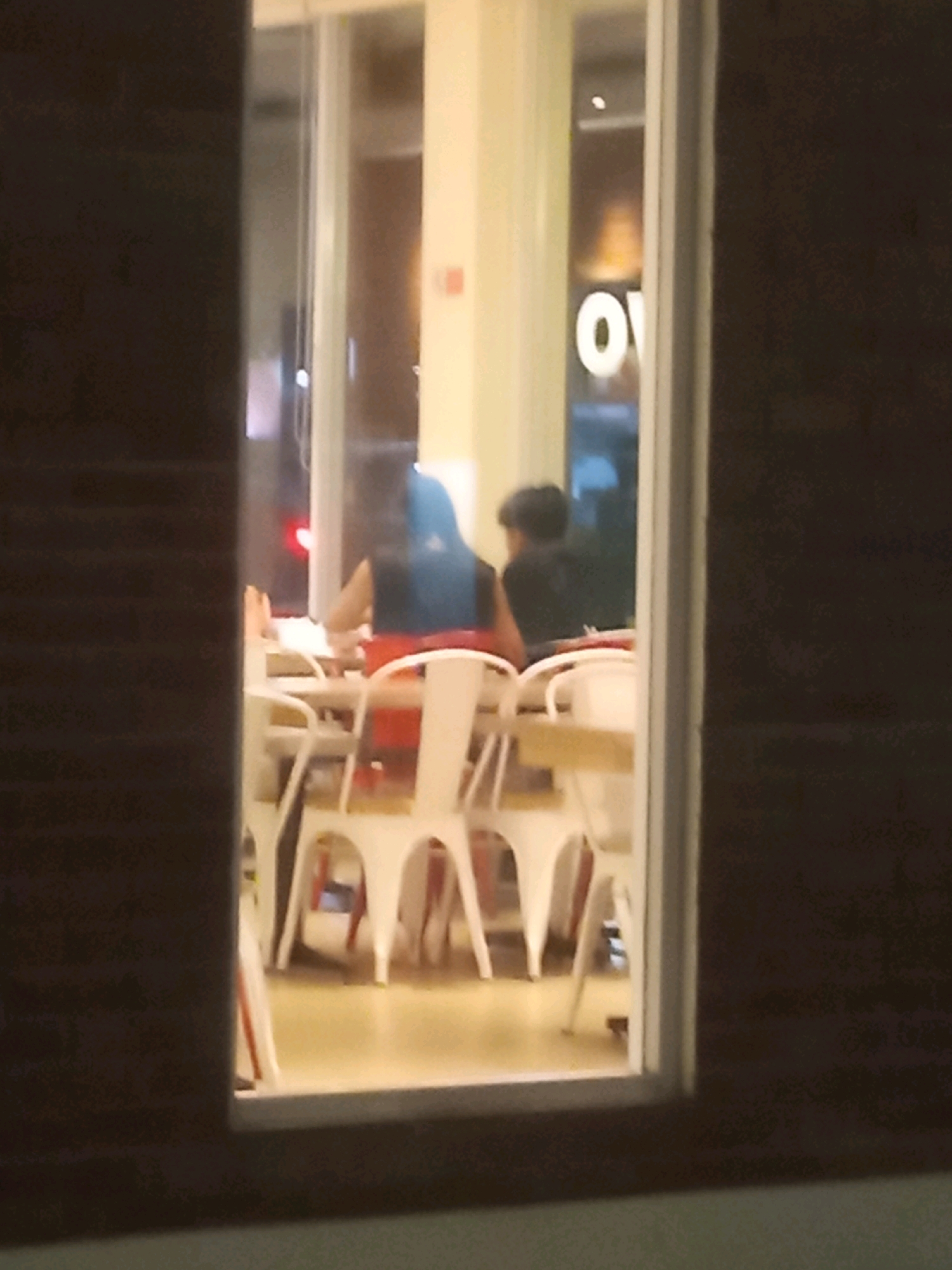 The image depicts the interior of a restaurant, viewed through a window from an exterior perspective. Two individuals are seated at a table, engaged in eating. The person on the left, with dark hair, is wearing an orange garment. The person on the right, also with dark hair, is wearing a dark shirt. White chairs surround the table. A portion of a white sign with the letters "OV" is visible on a wall in the background. The lighting suggests it is evening or night. The location is identified as Bontang, Indonesia.