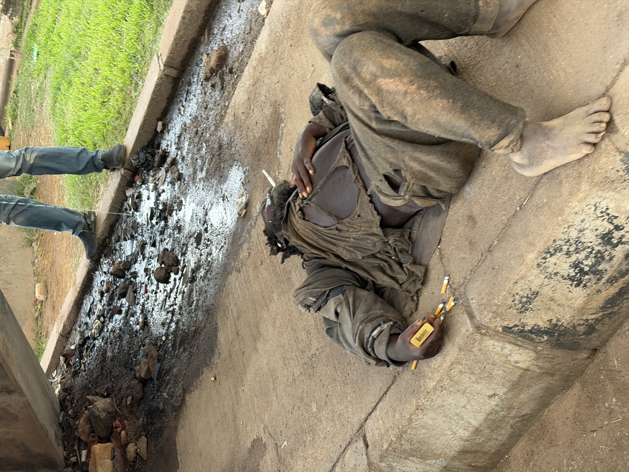 The image depicts a scene on a concrete surface next to a grassy area and a curb, with visible debris and water. In the foreground, a person is lying down, wearing tattered, dark clothing. Their torso is exposed, showing skin and torn fabric. One hand is resting near their chest, and in the other hand, they are holding several cigarette butts and a small, rectangular, gold-colored object with visible text that appears to say "WINCHESTER". The person's bare foot is visible on the right side of the frame. Their clothing appears dirty and worn, suggesting a state of disarray or destitution.
To the left of the person, the legs of another individual are visible, wearing blue jeans and dark shoes, standing near the curb. The area around the curb is filled with a mixture of dirt, rocks, and what appears to be refuse or mud. The grassy area to the left of the curb is bright green and appears well-maintained.
The lighting in the image suggests it is daytime, with shadows visible on the concrete. There are no explicit indicators of the weather, but the presence of water and mud suggests it may have recently rained or the area is prone to dampness. The scene appears to be outdoors, possibly in an urban or semi-urban environment given the concrete and curb. The overall impression is one of neglect and hardship. The text "WINCHESTER" on the gold object is the only visible text. The image was taken in Gura Top, Nigeria.