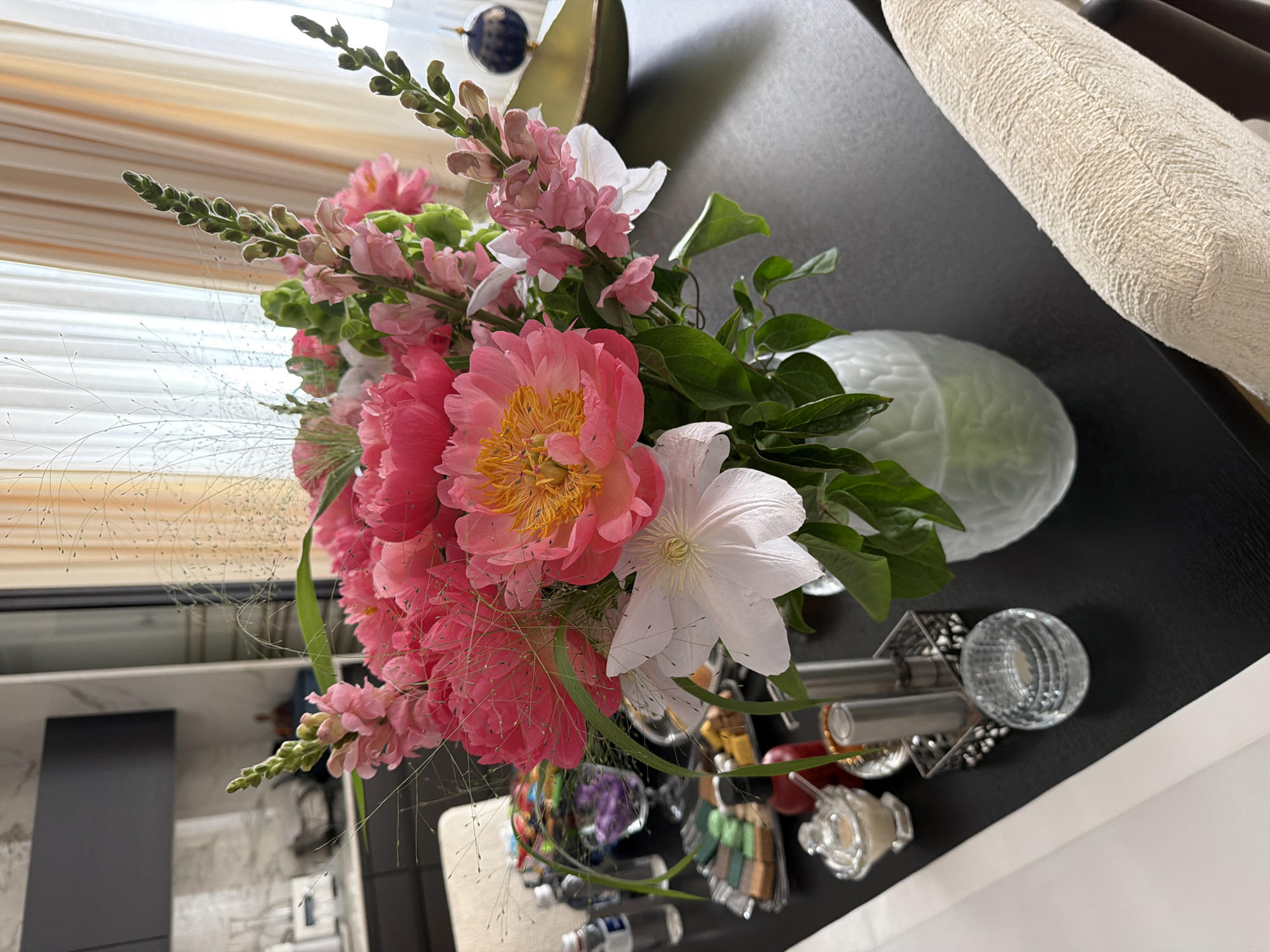 A bouquet of pink peonies and snapdragons sits in a textured glass vase on a black table. The flowers are arranged with delicate grasses and greenery. In the background, sheer curtains are visible. On the table, there are several decorative items, including a silver tray with colorful candies, a small glass bowl with a lid, a metal napkin holder, and a clear glass votive holder. A chair upholstered in textured cream fabric is positioned to the right of the table. The setting appears to be an indoor dining or living area.
