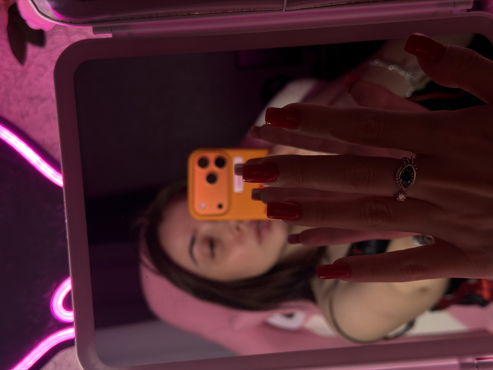 A person is reflected in a mirror, taking a selfie with an orange phone. The person has long, red acrylic nails and is wearing a ring on their left hand. They are seated in a pink chair, and the background is illuminated by pink neon lights, shaped like cat ears.
