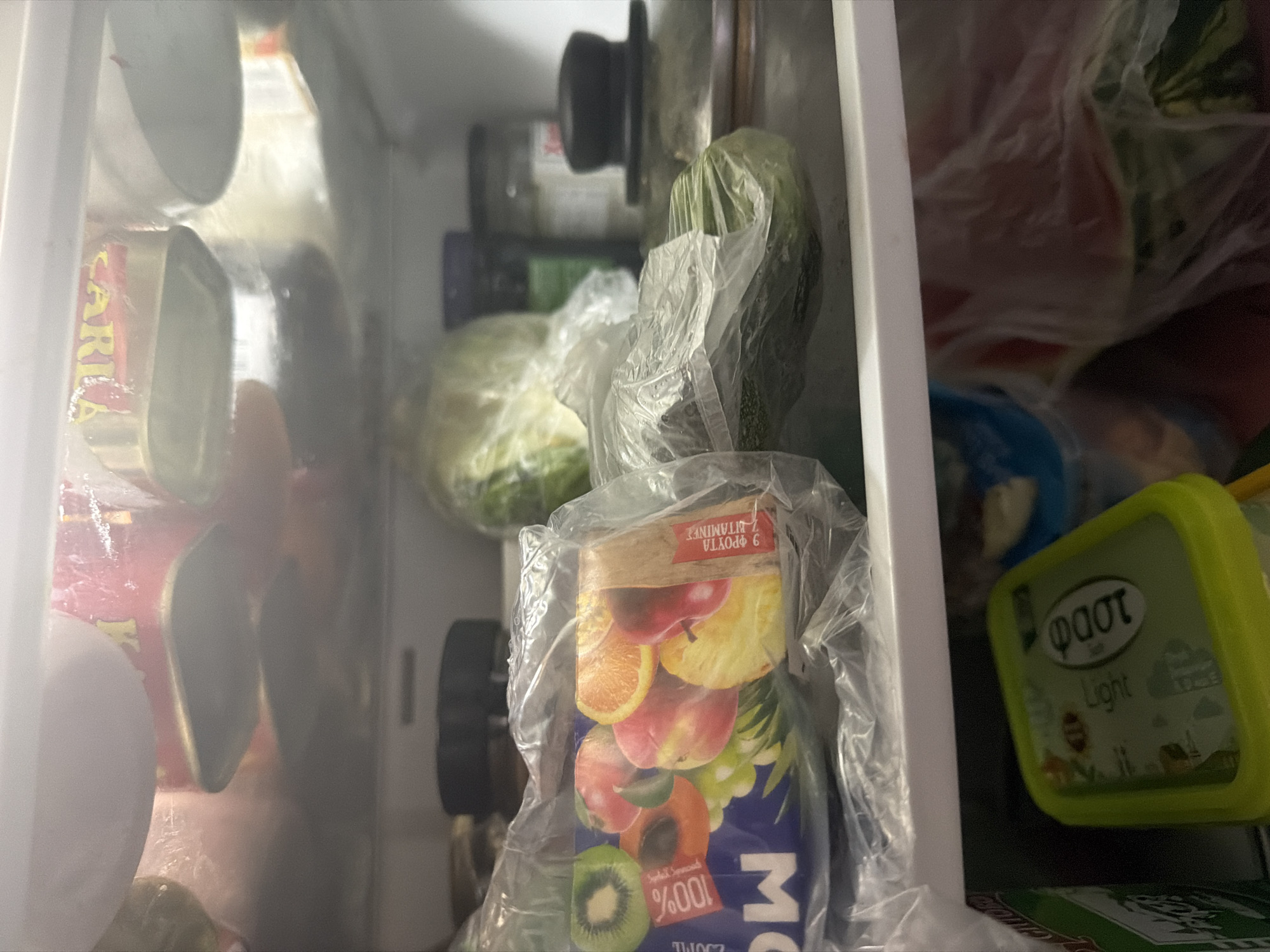 The image shows the inside of a refrigerator containing various food items. On the top shelf, there are some canned goods with labels visible. The middle shelf holds a carton of juice with images of fruits, a head of lettuce or cabbage, and a zucchini wrapped in plastic. There are also some jars and containers on this shelf. The bottom shelf contains a green tub of what appears to be yogurt or a dairy product with text in Greek, and a partially visible watermelon in a plastic bag. The refrigerator is located in Dromolaxia - Meneou Municipality, Cyprus.