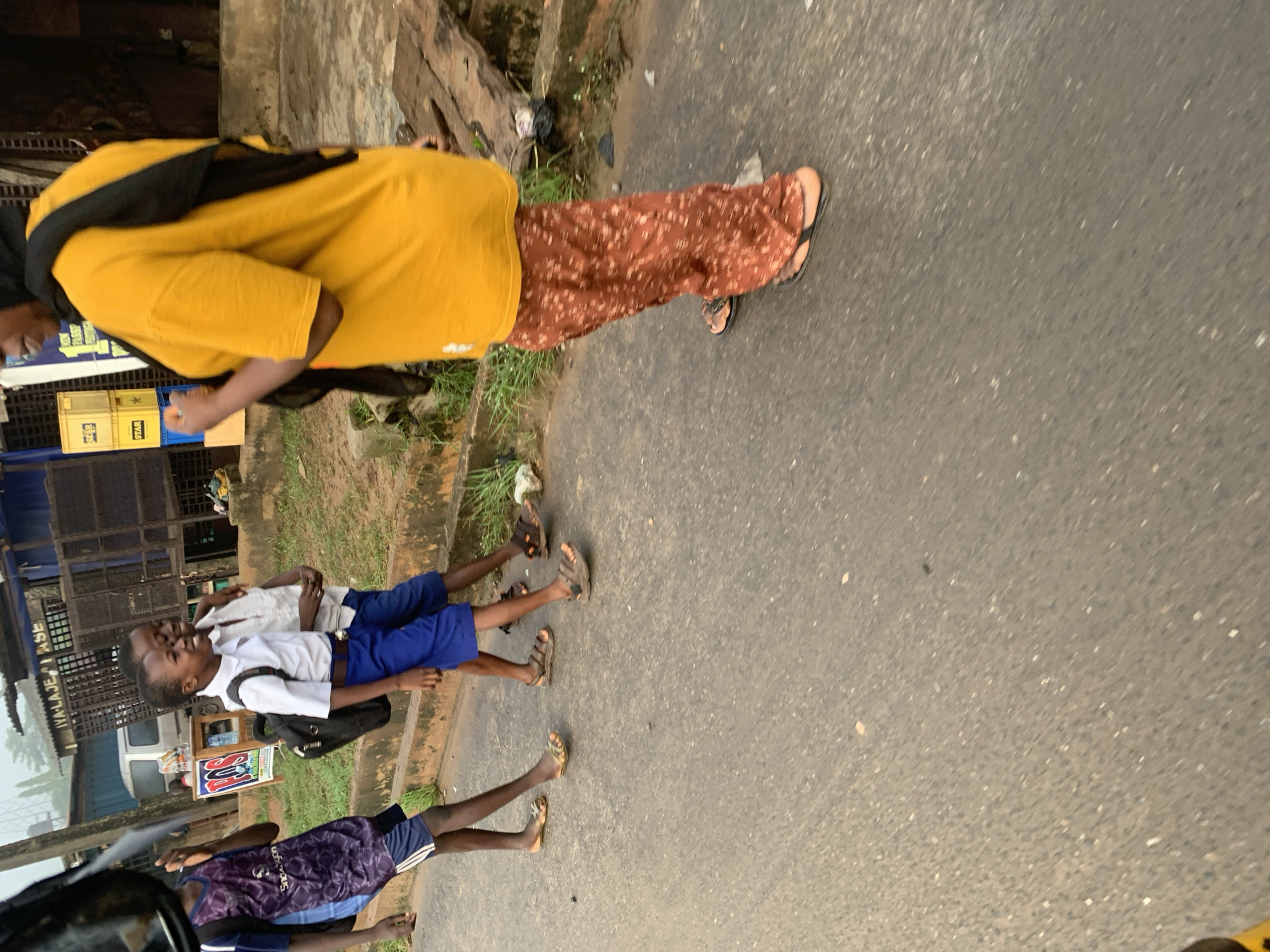 The image shows a group of people on a paved road, likely in Shagamu, Nigeria. In the foreground, a person wearing a bright yellow t-shirt and patterned brown pants with a black bag draped over their shoulder is walking away from the camera. Behind them, three young boys are standing. Two of the boys appear to be wearing school uniforms, one with a white shirt and blue shorts, and the other with a white shirt and black shorts. They are smiling and interacting with each other. The third boy is wearing a purple and blue athletic outfit. The background features a building with signage and some greenery, suggesting an urban or semi-urban environment. The lighting indicates it could be daytime, and the overcast conditions hint at cloudy weather. The scene appears casual, with the people dressed in everyday clothing and footwear.