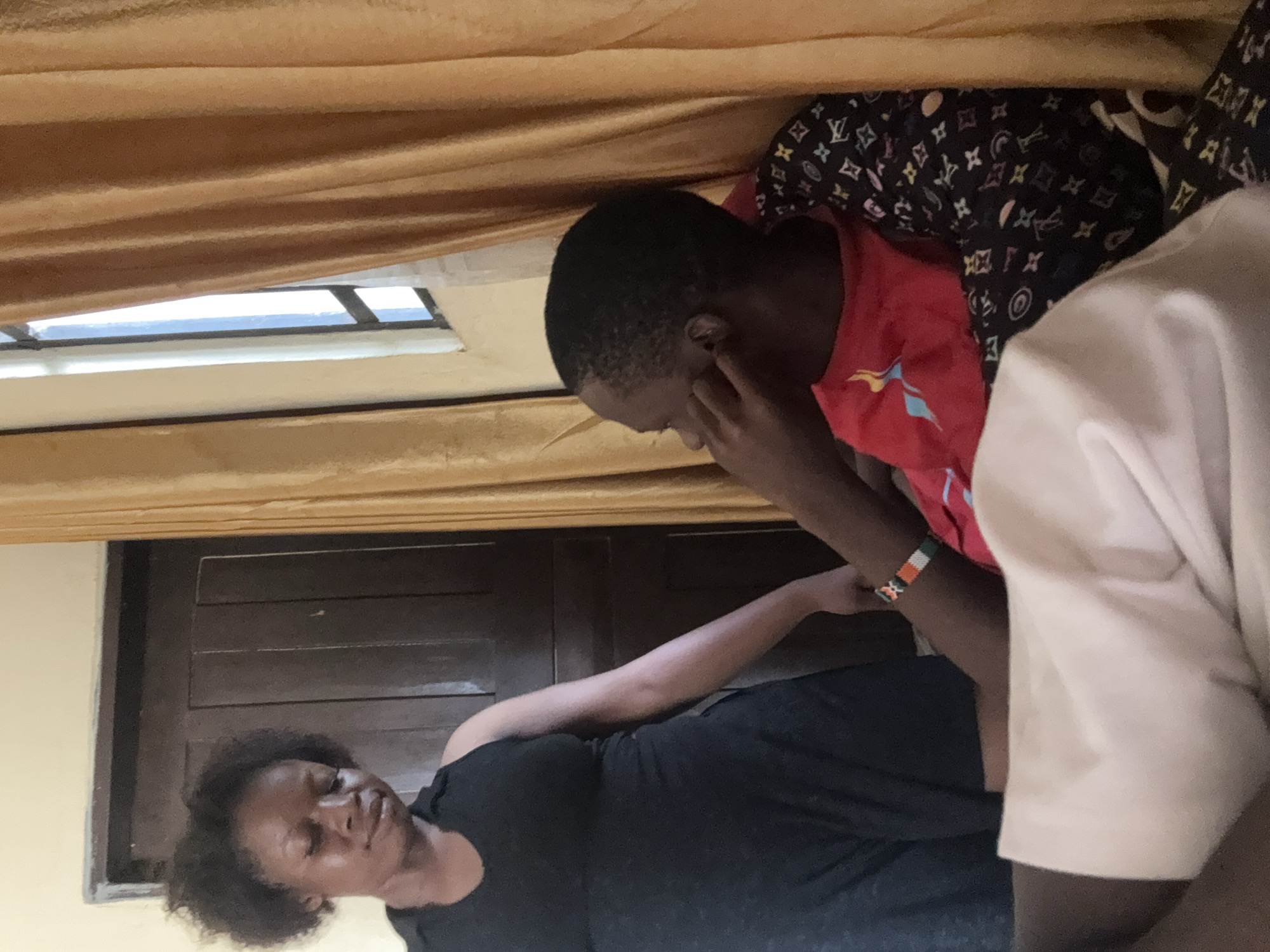 An indoor scene, captured in Ikot Ekpene, Nigeria, features two main individuals and a partial view of a third. The image is oriented such that the subjects appear to be lying on their sides. In the lower left, a woman with dark, curly hair and a black top is positioned horizontally, appearing to be resting with her eyes closed or partially closed. Her left arm extends towards the center.
Above and to her right, a young person with short dark hair, wearing a red short-sleeved shirt with a light blue and yellow graphic, is seen from the side, leaning over the woman. Their right hand is near their face, while their left arm extends towards the woman's arm, possibly in a comforting or supportive gesture. A colorful beaded bracelet, featuring orange, white, green, and black sections, is visible on the young person's left wrist. Behind this individual, a dark fabric with a white patterned design, resembling the Louis Vuitton monogram (showing "LV" initials and floral motifs), is visible.
Partially seen on the far right is another individual wearing a light-colored (likely white or cream) top. In the background, dark brown wooden paneling or a door with horizontal slats is present behind the woman. The upper part of the scene is framed by pleated brown curtains, which partially obscure a window or vent through which a bright exterior light, likely the sky, is visible. The atmosphere suggests an intimate moment, possibly of rest or supportive interaction.