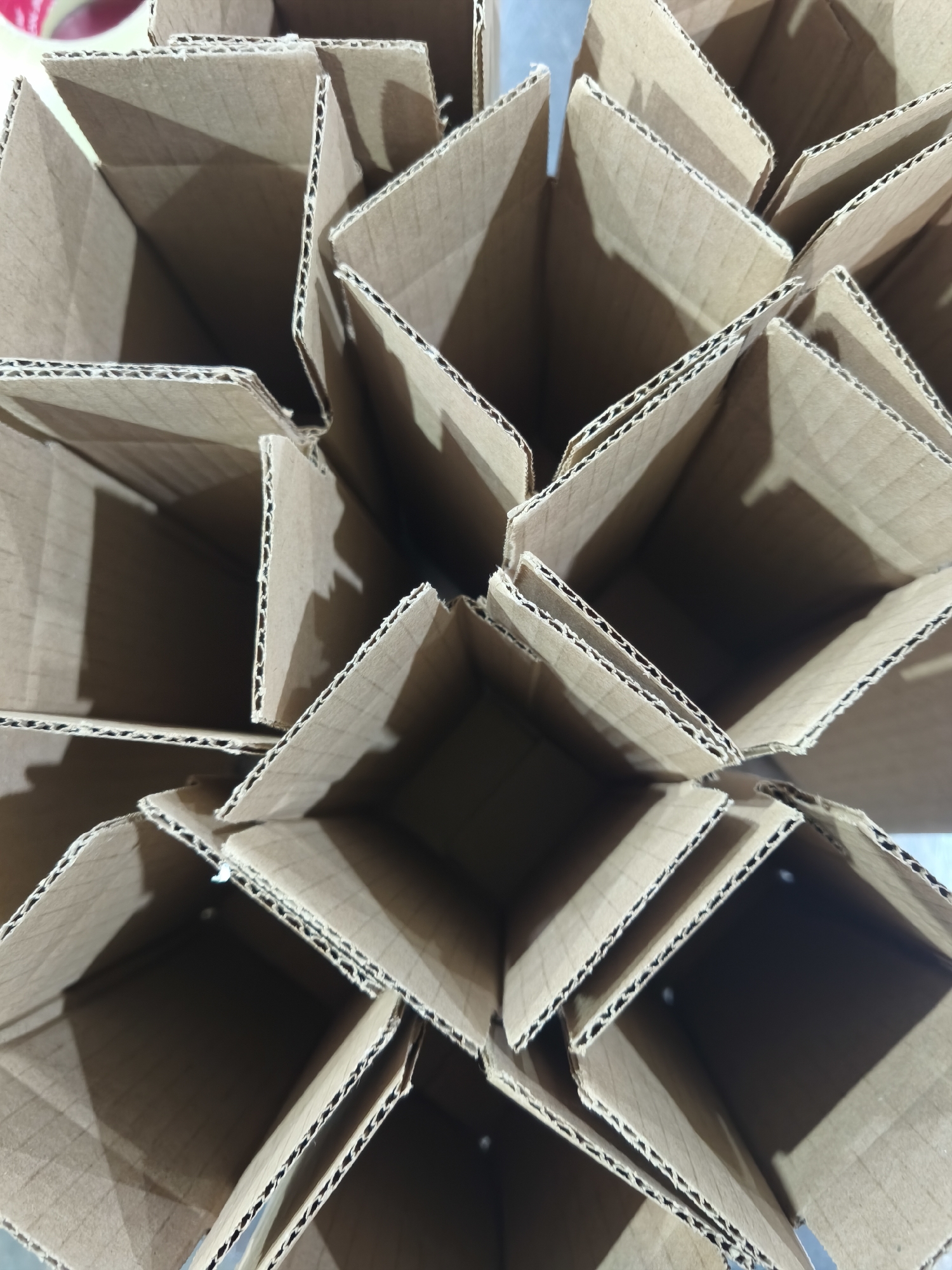 A high-angle, close-up photograph displays multiple unfolded cardboard boxes arranged in a stack, creating a geometric pattern of diagonal lines and square openings. The boxes appear to be made of corrugated cardboard. The location is not discernible from the image.