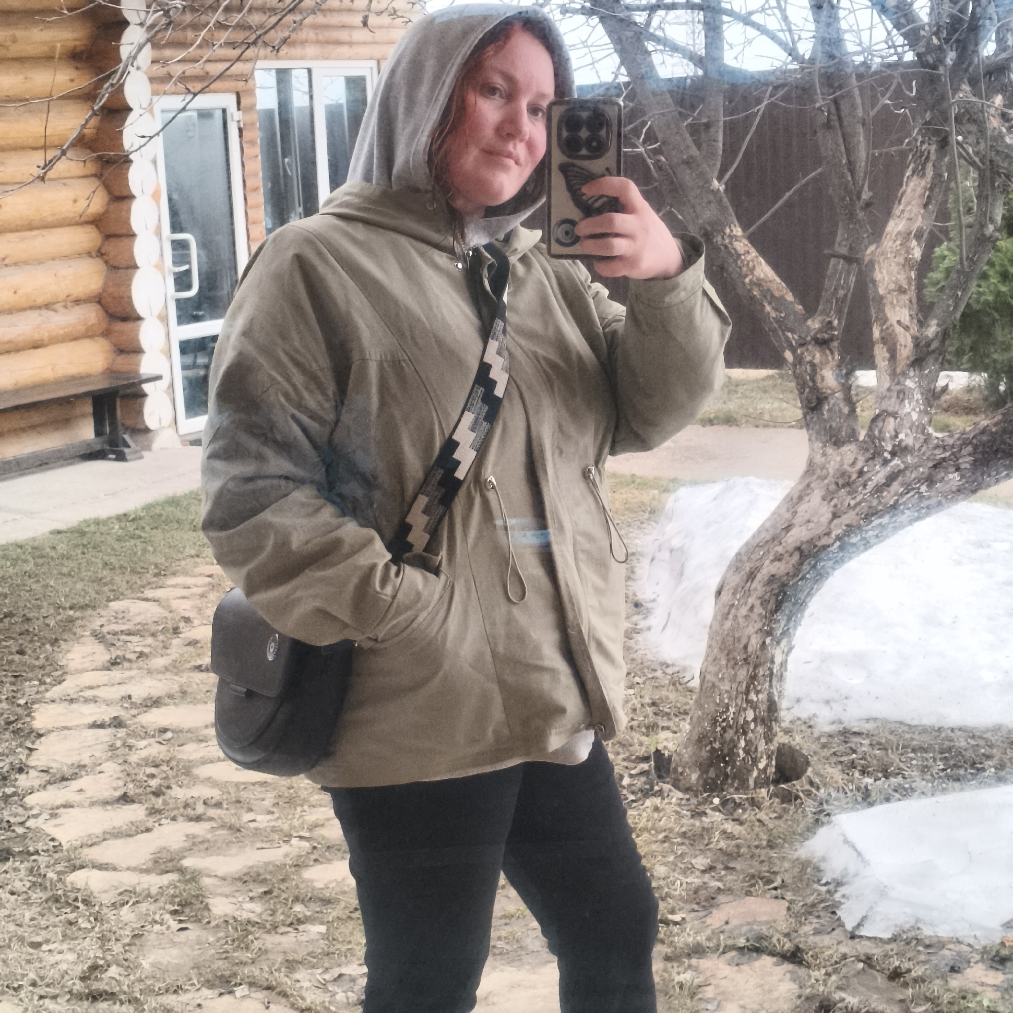 A woman is taking a selfie in an outdoor setting in the city of Chaikovsky, Russia. She is wearing a hooded jacket, dark pants, and a crossbody bag. The location appears to be a yard with a wooden log building in the background and patches of snow on the ground. A tree is visible to the right of the woman. The ground is covered with paving stones and sparse vegetation.