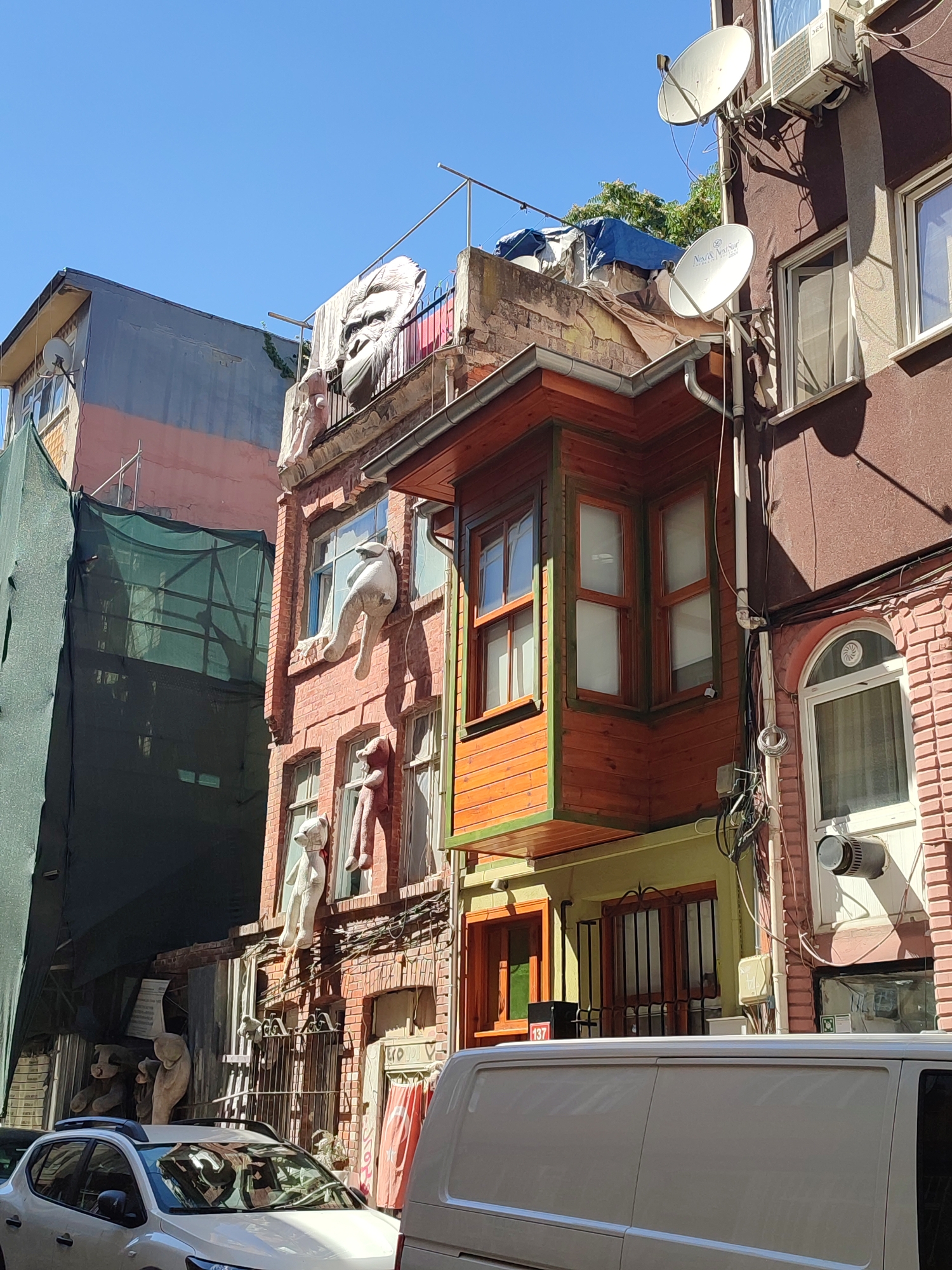 The image depicts a narrow street in Kadıköy, Turkey, characterized by multi-story buildings with varying architectural styles and facades. On the left, a building is covered with green scaffolding netting, obscuring its appearance. To its right, a brick building features several white, humanoid figures, appearing to be sculptures or artistic installations, draped over windows and balconies. One figure, resembling a large ape's head, is visible on the rooftop.
The central building is a narrow, multi-story structure with prominent wooden bay windows painted in orange and green. Below these windows, the facade is a mix of brick and what appears to be a pale yellow stucco. Iron gates are visible at street level, with some decorative elements.
To the right of the wooden-accented building is another structure with a darker, reddish-brown facade. This building has multiple windows with white frames, an air conditioning unit mounted high up, and a satellite dish attached to the exterior wall. A white van is parked in the foreground, partially obscuring the lower parts of the buildings. A white car is also visible on the left side of the frame. The sky is clear and blue, indicating a sunny day. The overall impression is one of a densely built urban environment with eclectic and unusual decorations on display.