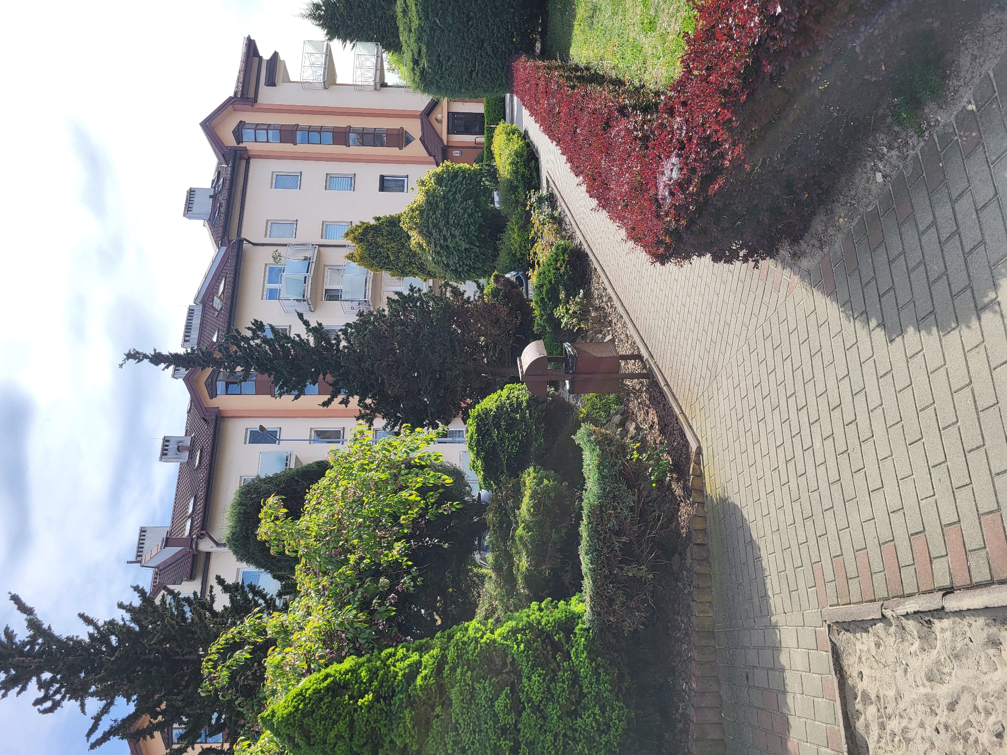 The image depicts an exterior view of a multi-story building in an urban setting. The building is painted in light beige tones with brown roofing and trim. It has multiple windows and balconies. Lush green landscaping and bushes surround the base of the building. A pathway made of interlocking bricks runs parallel to the building, bordered by a garden bed with red shrubbery. The sky is partly cloudy with hints of blue. The perspective is from a slightly low angle, emphasizing the height of the building and the depth of the pathway.