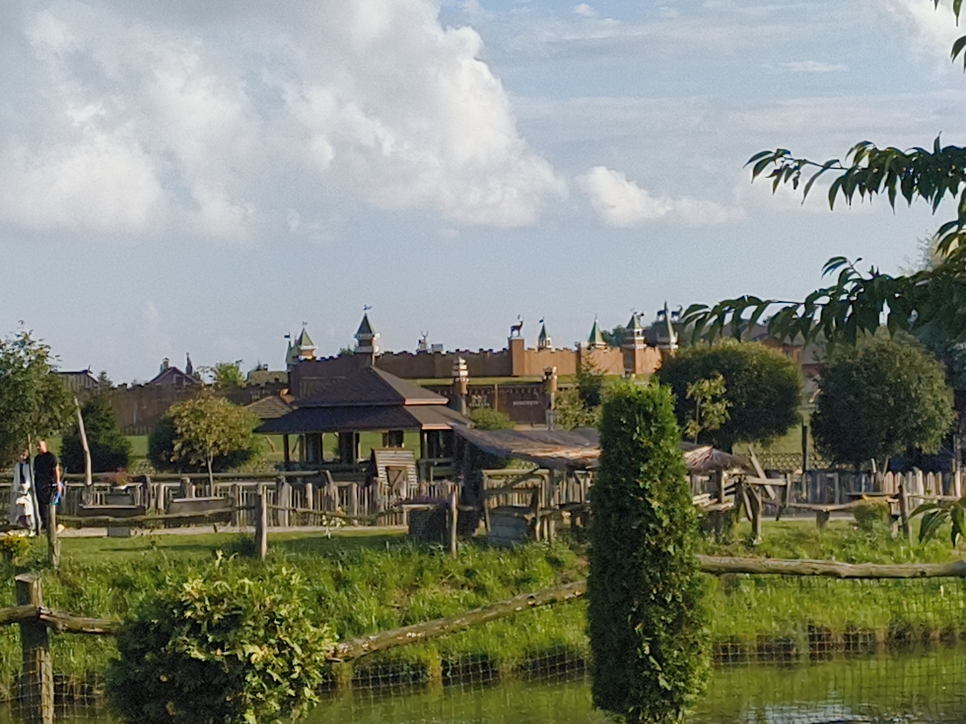 A picturesque rural landscape is depicted, featuring a body of water in the foreground. Beyond the water, a wooden fence separates the immediate area from a grassy expanse. In the mid-ground, various wooden structures, including a covered pavilion and enclosures, are visible, suggesting a park or attraction. Behind these structures, a large, castle-like building with towers and ornate details dominates the background, hinting at a themed environment. Lush green trees and foliage are scattered throughout the scene, with a tall, slender evergreen tree standing prominently on the right side. The sky is partly cloudy, with soft white clouds against a pale blue backdrop. A person in dark clothing stands near the left edge of the mid-ground, interacting with another figure in white. The overall atmosphere is serene and evokes a sense of a recreational or entertainment venue in a natural setting.