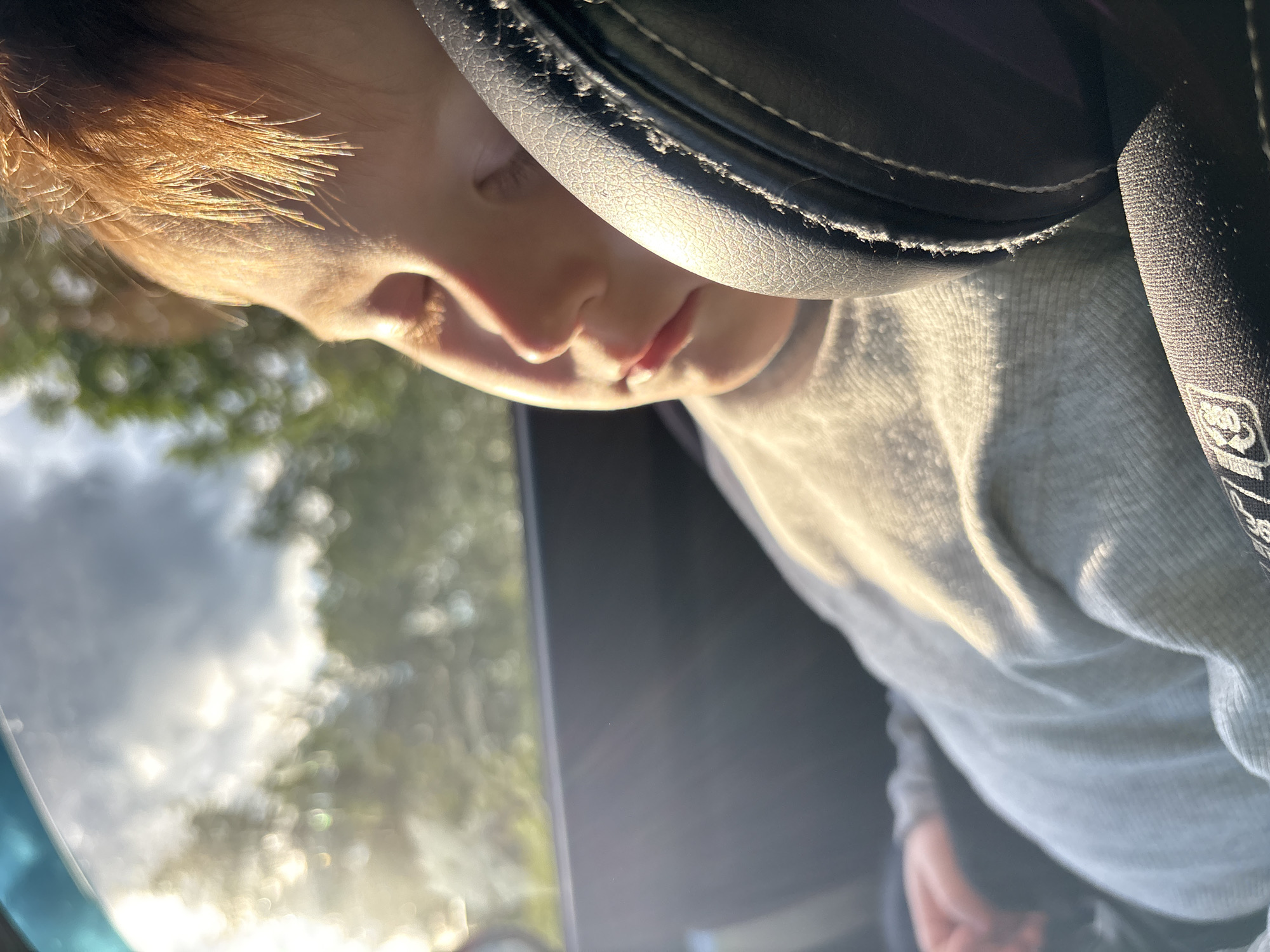 A child is inside a car, likely asleep. The child has reddish-blonde hair and is wearing a grey long-sleeved shirt. The head is close to the car's interior, specifically the headrest of a seat. Outside the car window, there are blurred shapes suggesting trees and sky. The light is bright, indicating daylight. No details suggest a location in Izhevsk, Russia.